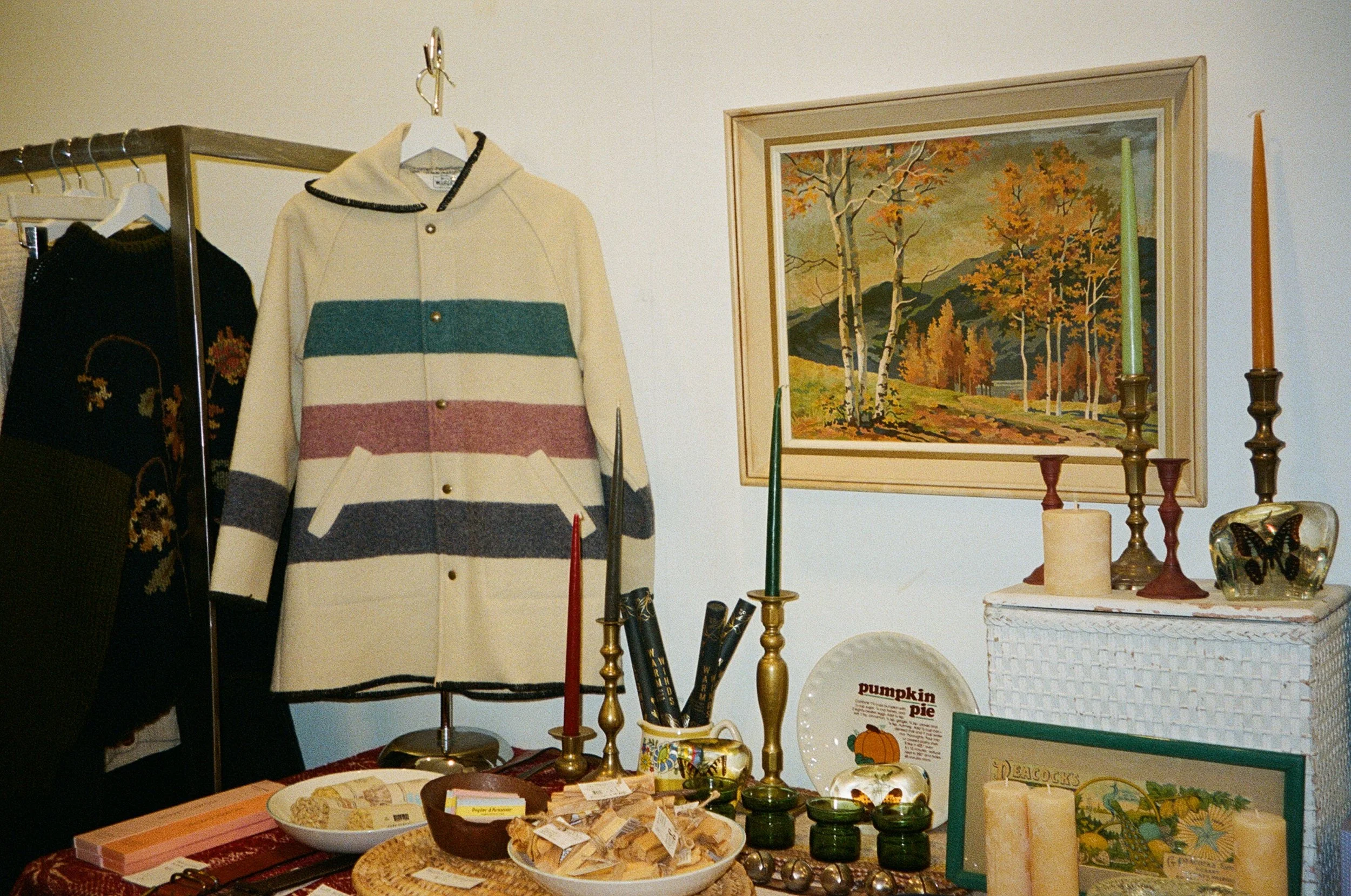 Clothing display with a striped coat, a black sweater with floral embroidery, and various home decor items including candle holders, candles, a decorative plate with pumpkin pie, and picture frames, set against a white wall with a landscape painting.