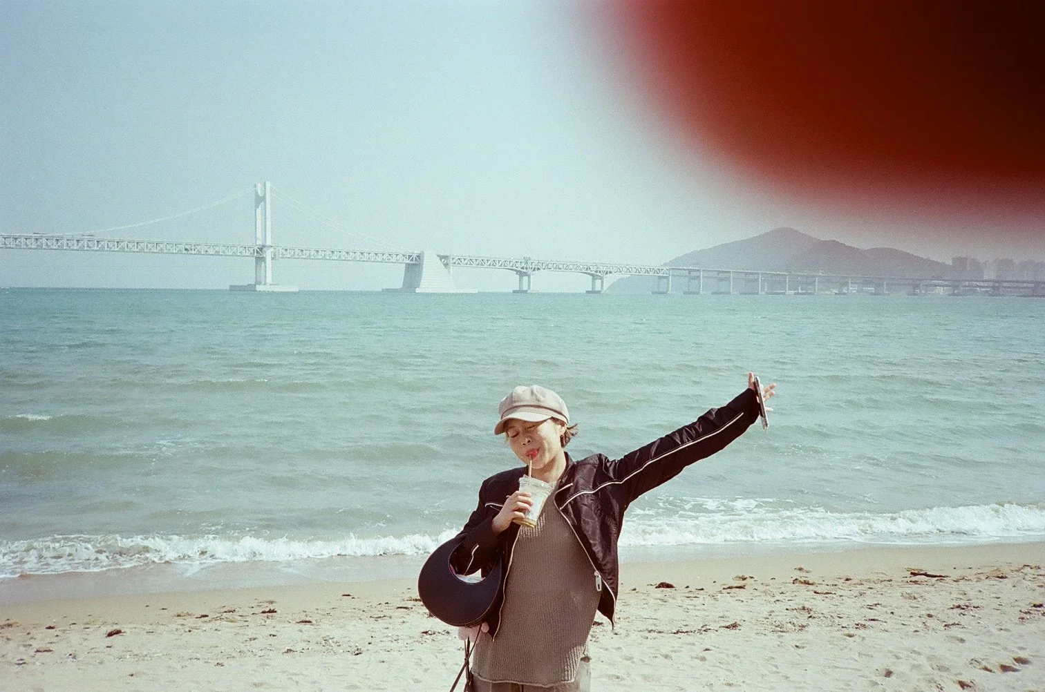 A woman standing on a sandy beach, holding a drink and a guitar, with her arms outstretched and eyes closed, in front of the ocean and a bridge in the distance.