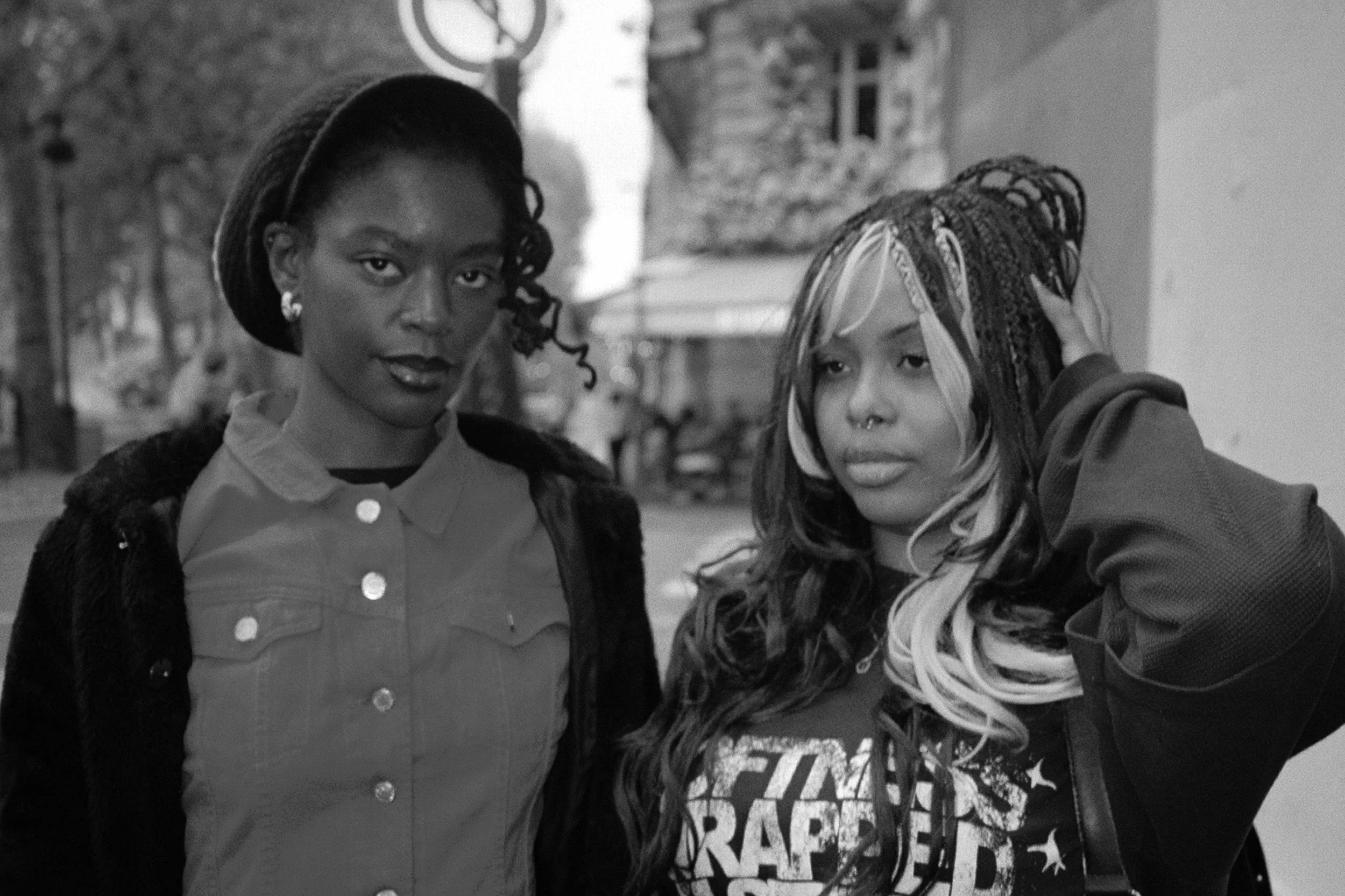 Two women with alternate hairstyles, one with dark curly hair and the other with long hair with lighter streaks and braids, standing outdoors near a wall and a security sign. The woman on the left wears a button-up shirt and earrings, while the woman on the right is touching her hair and wears a hoodie.