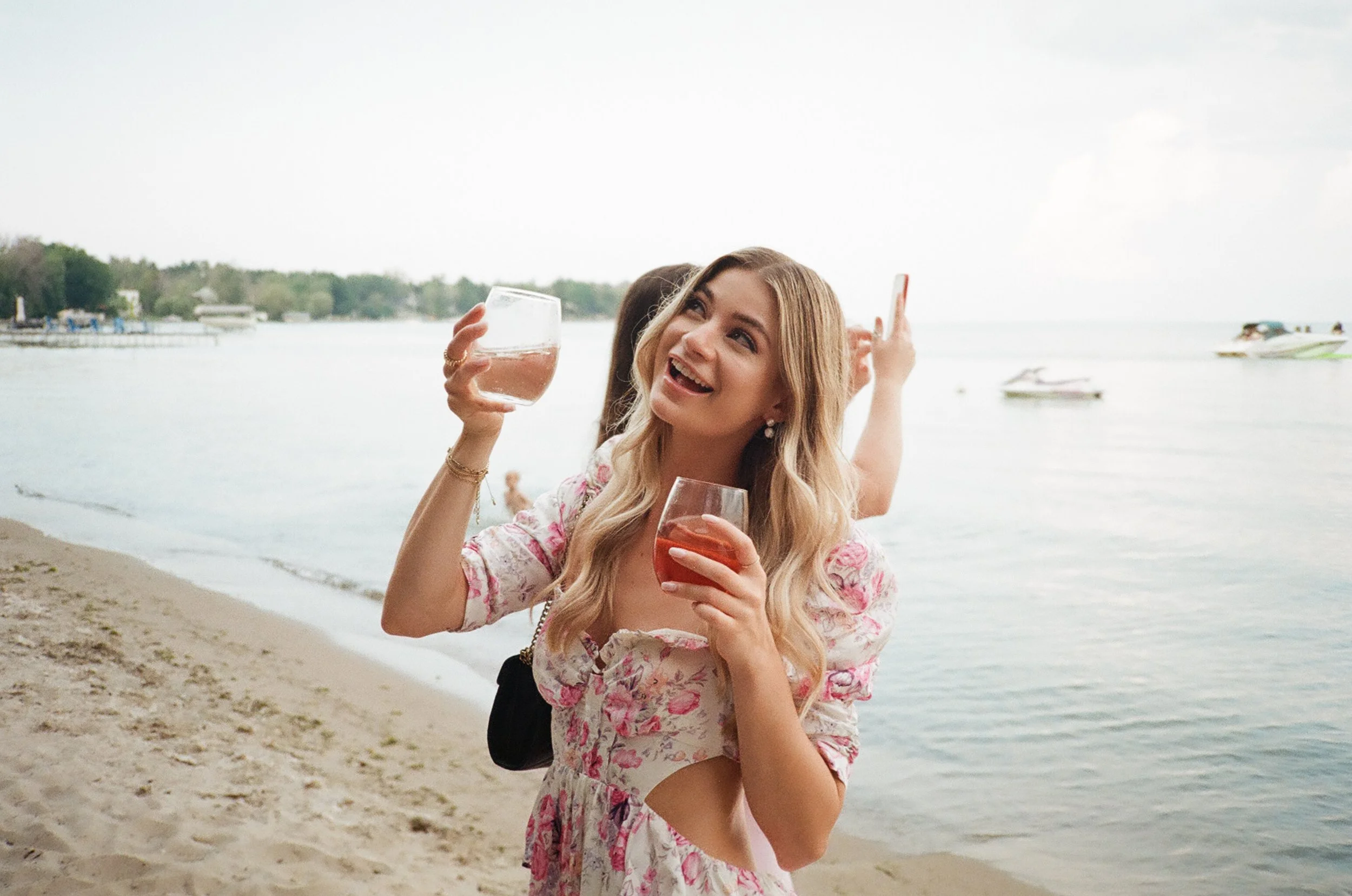 A young woman in a floral dress holding a glass of wine and smiling on a beach with water and boats in the background.
