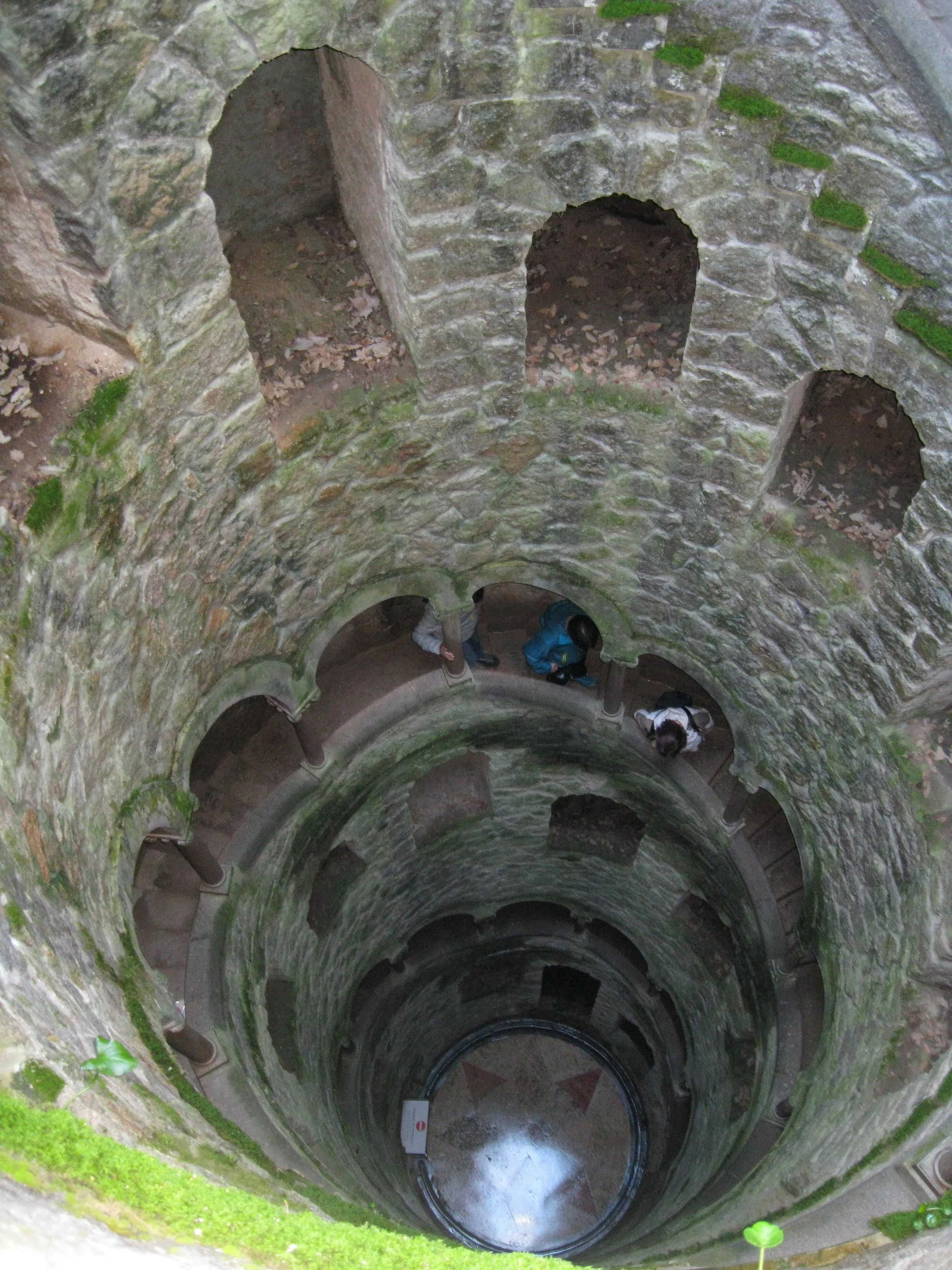 View looking down into a stone well with multiple arches and a circular bottom, with people standing on the edge.
