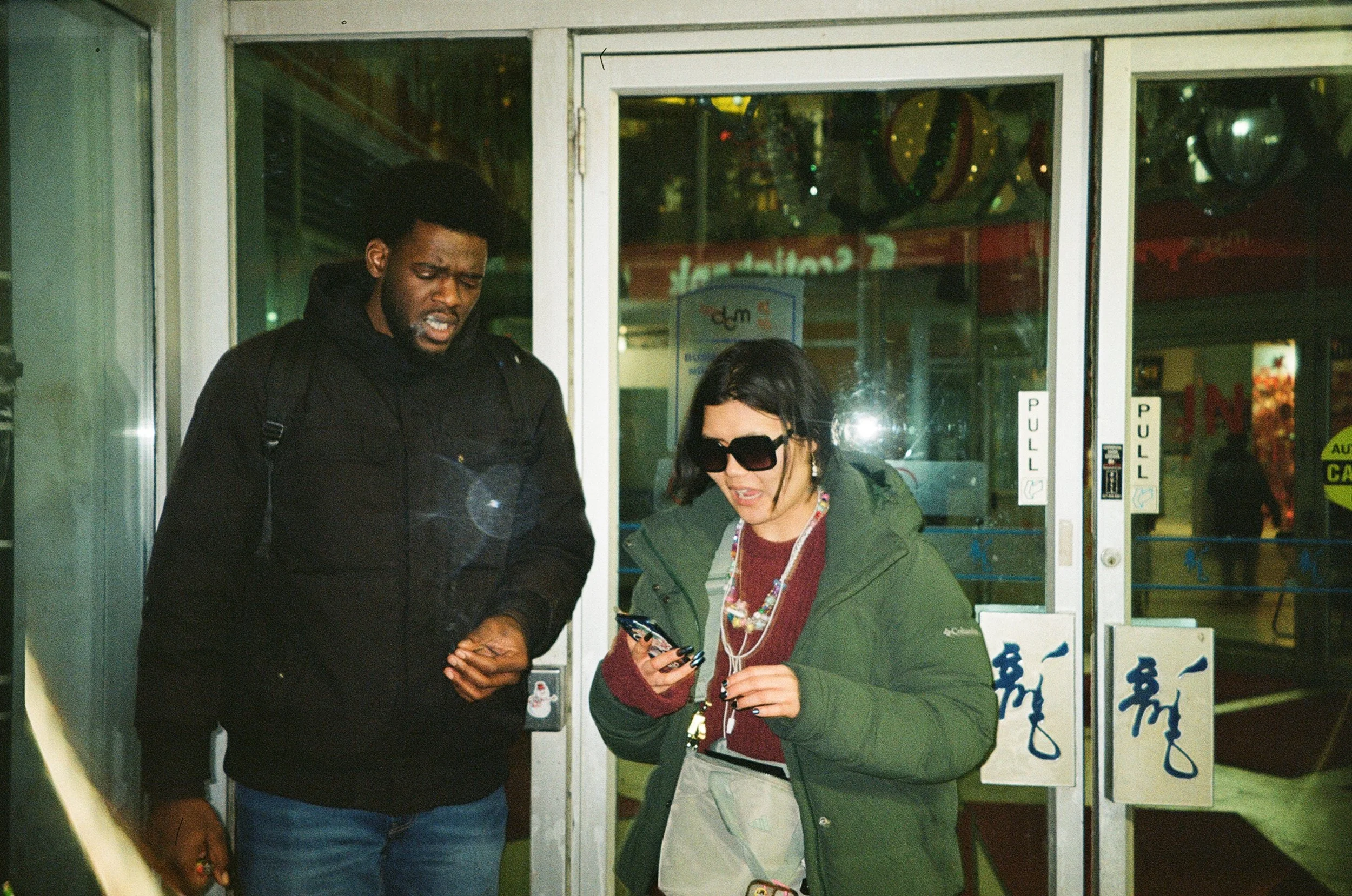 A man and woman standing outside a store entrance, looking at a smartphone, woman wearing large sunglasses and green jacket, man in black jacket, with reflective glass doors behind them.