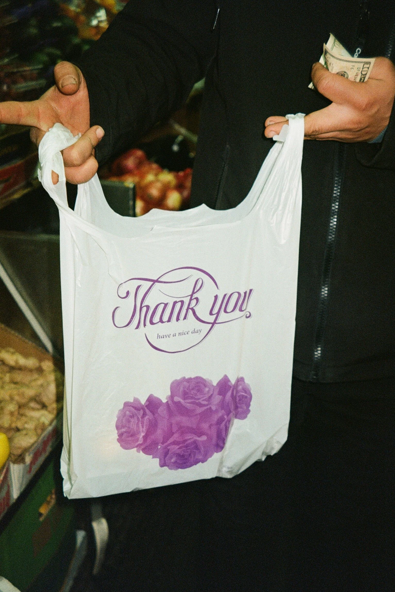 A person holding a white plastic grocery bag with purple roses and thank you message printed on it, while also holding cash. The person is wearing a black jacket.