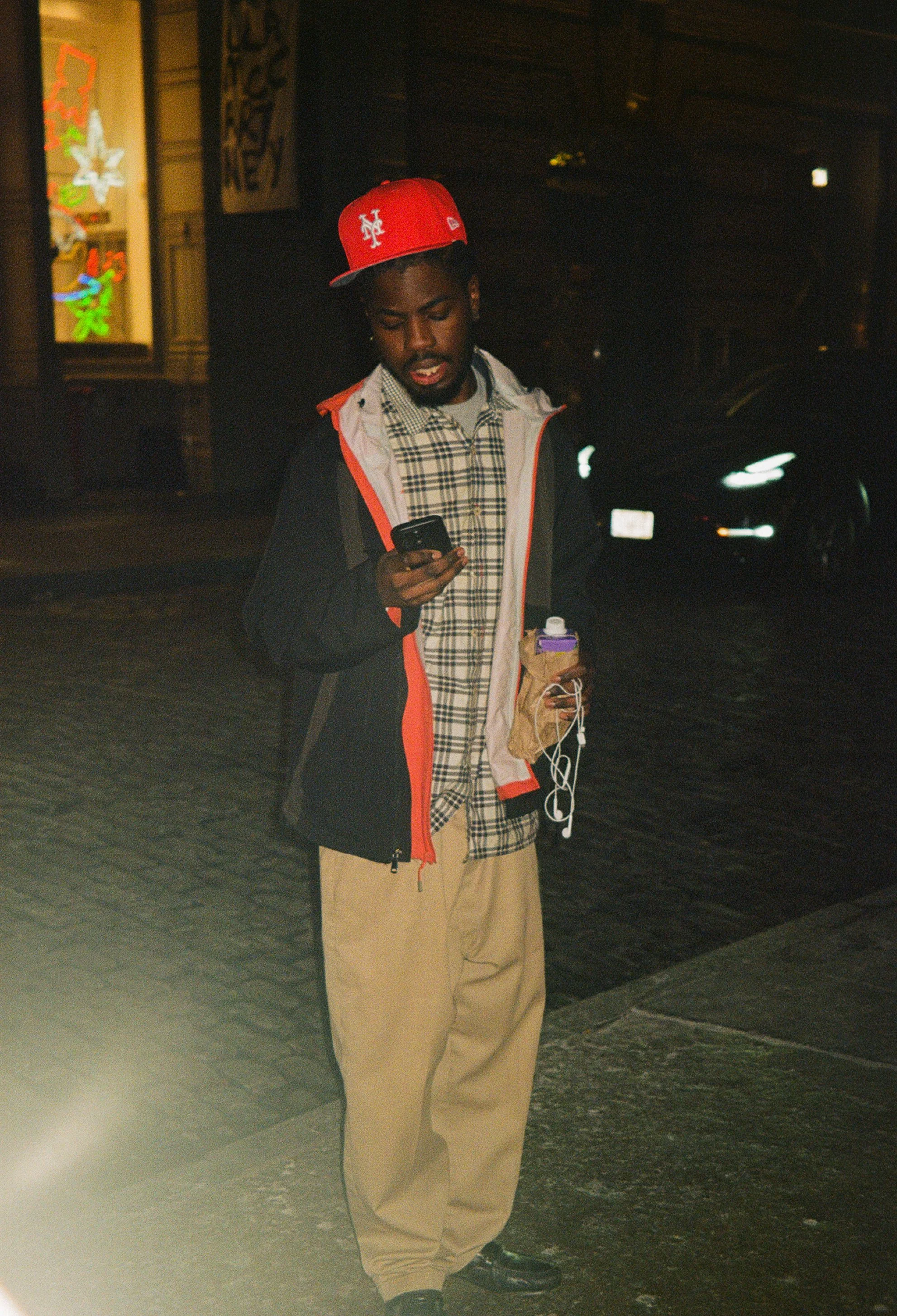 A young man standing outdoors at night, looking at his phone, wearing a red cap, checked shirt, beige pants, and a black jacket with red accents. He is holding a brown paper bag with a purple water bottle and earphones.