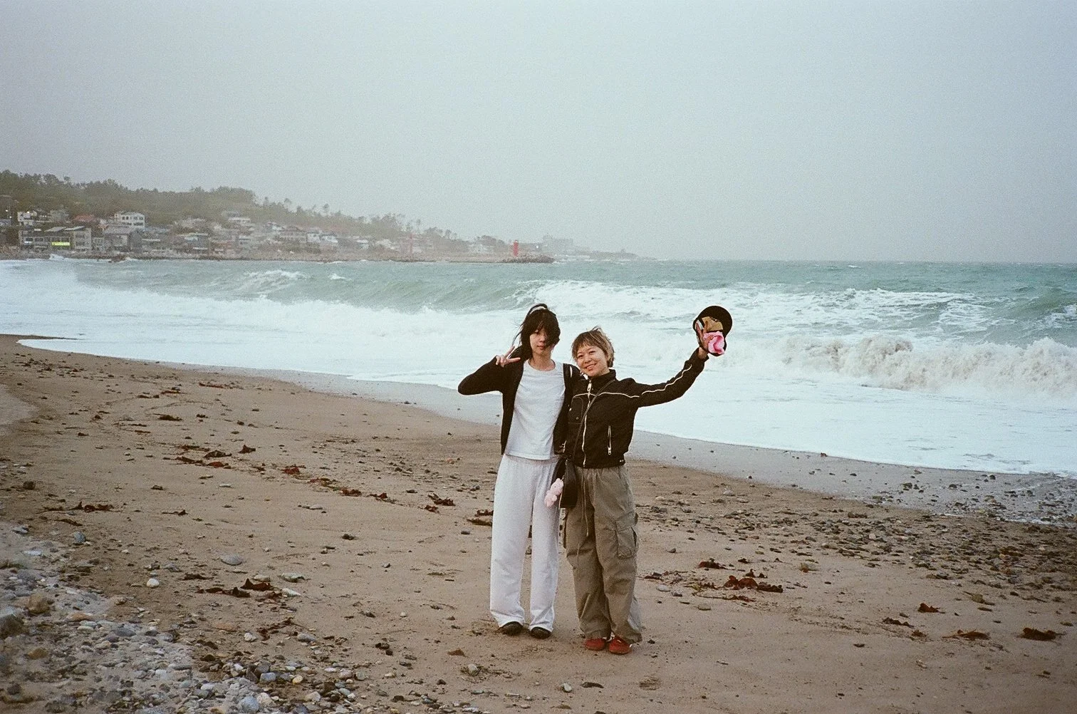 Two women standing on a beach near the ocean, one holding a small purse and the other making a peace sign next to her, with waves crashing in the background.