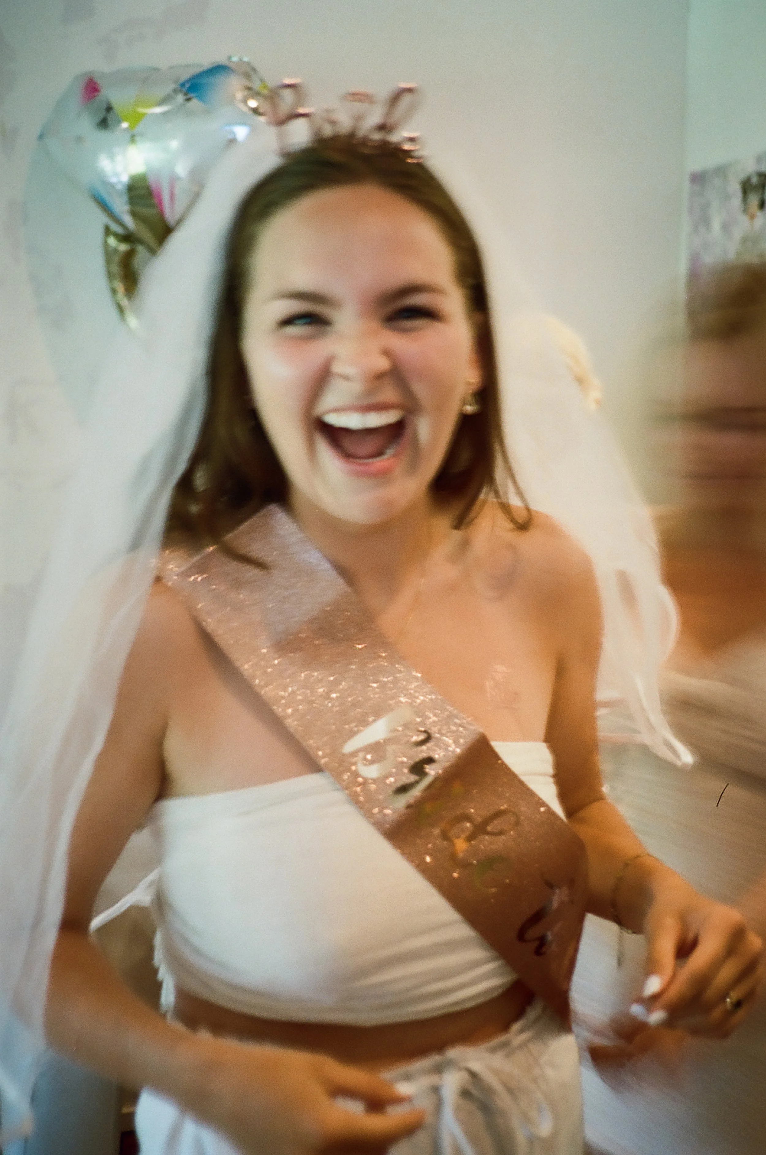 A woman with a crown and a glittery birthday sash is celebrating her birthday, smiling and laughing excitedly, with a birthday balloon in her hair.