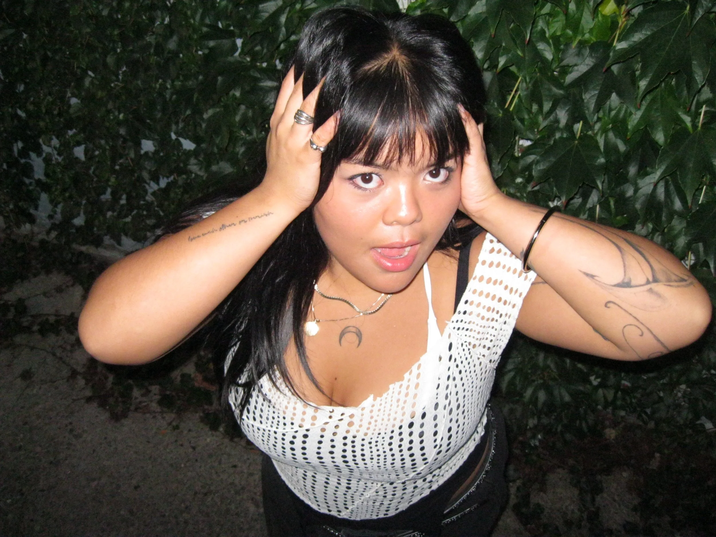 A woman with black hair and multiple tattoos, wearing a white mesh top and jewelry, poses with her hands on her head in front of green leafy bushes.