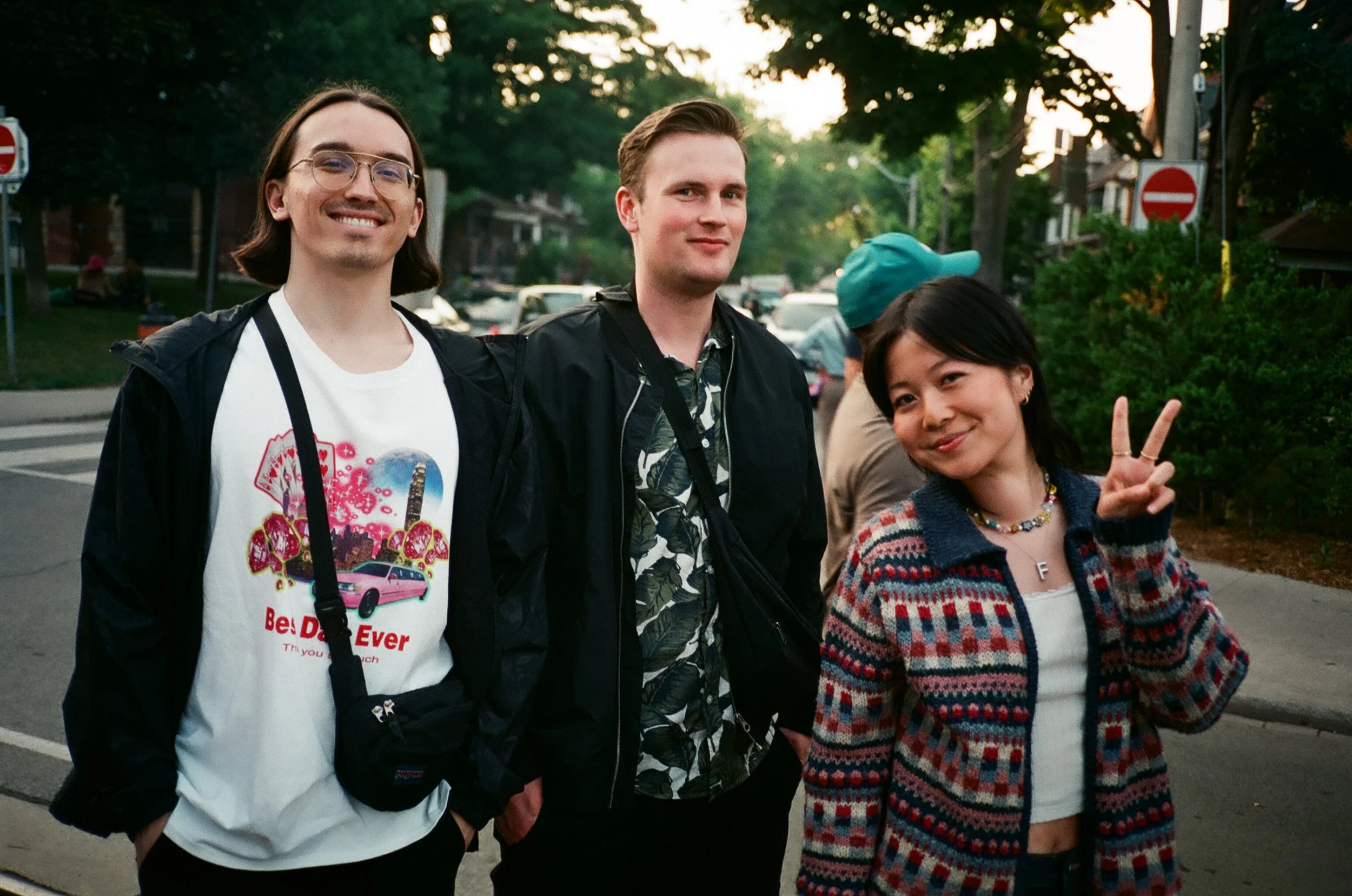 Three friends standing on a sidewalk in a residential neighborhood during sunset, smiling at the camera. The person on the left is a young man with glasses, wearing a white t-shirt and a black jacket. The person in the middle is a young man with shor