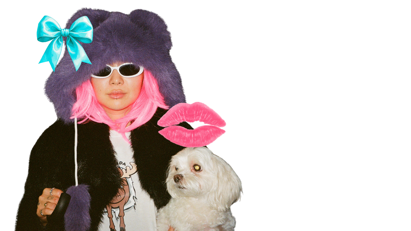 Person with pink hair, wearing sunglasses and a fuzzy hat with a blue bow, holding a small white dog, with pink lips and a black coat.
