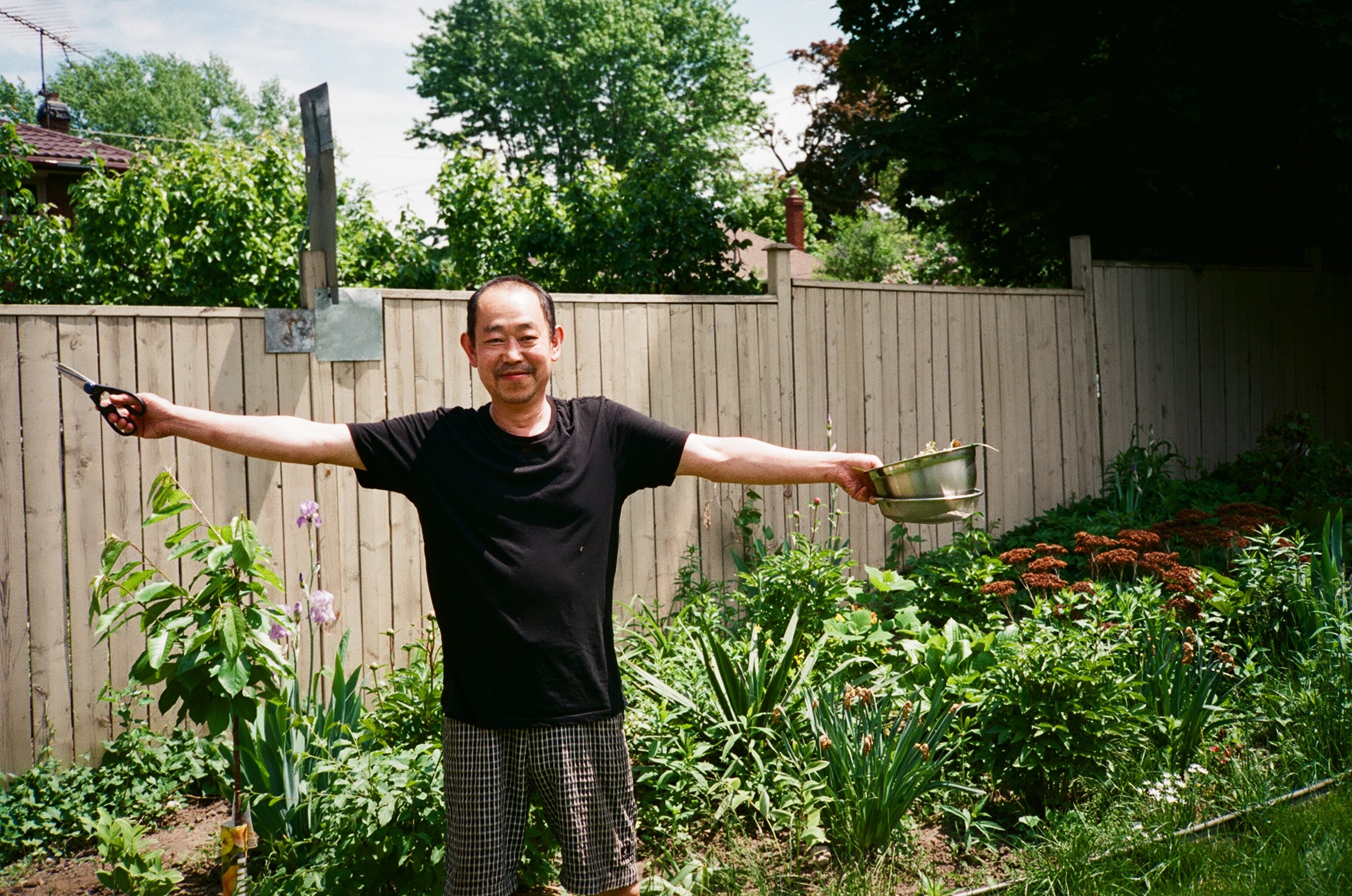 A man in a black t-shirt and checkered shorts standing in a backyard garden with arms outstretched, holding gardening tools, with a wooden fence and green foliage in the background.