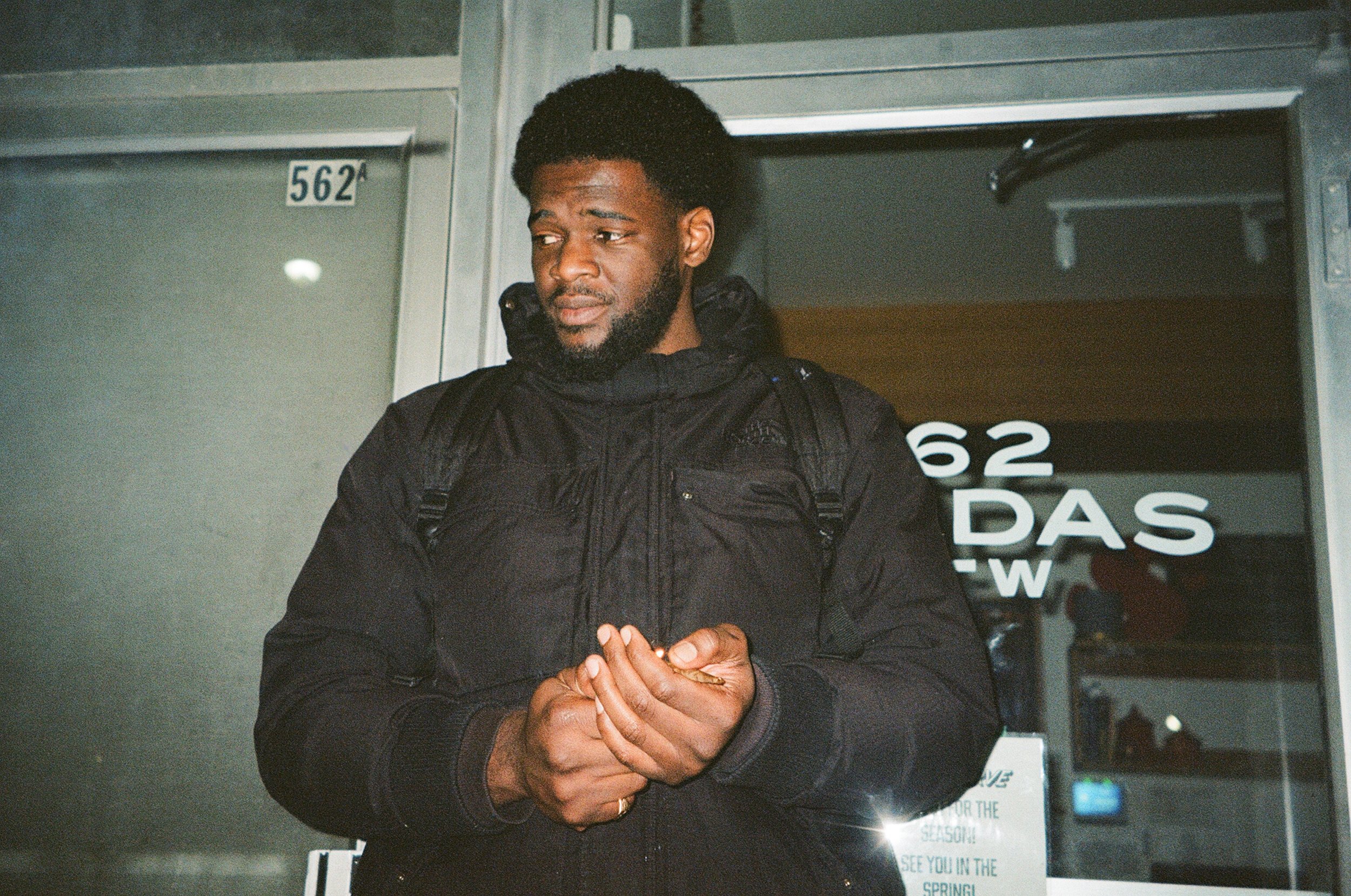 A man with a beard and short curly hair wearing a black jacket and backpack, standing outside a building, appears to be awaiting or in thought.