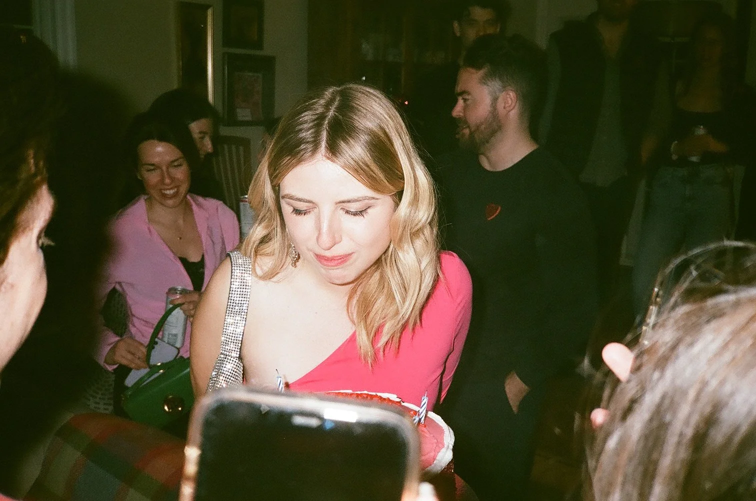 A blonde woman in a pink dress is surrounded by people at a party, holding a birthday cake with lit candles, with several people smiling and gathering around her in a dimly lit room.
