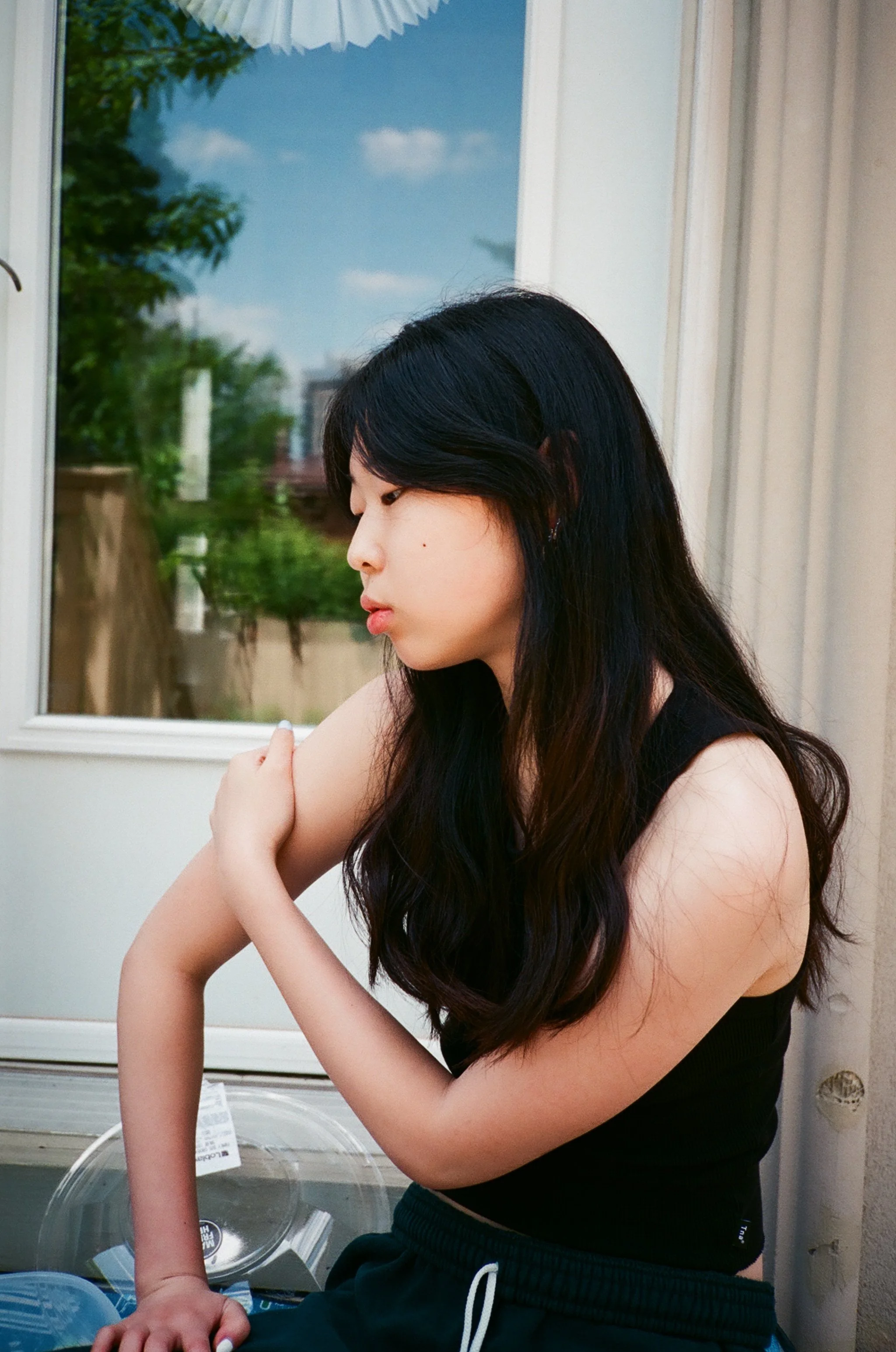 A young woman with long black hair sitting near a window, wearing a black sleeveless top and dark pants, looking down and touching her arm.