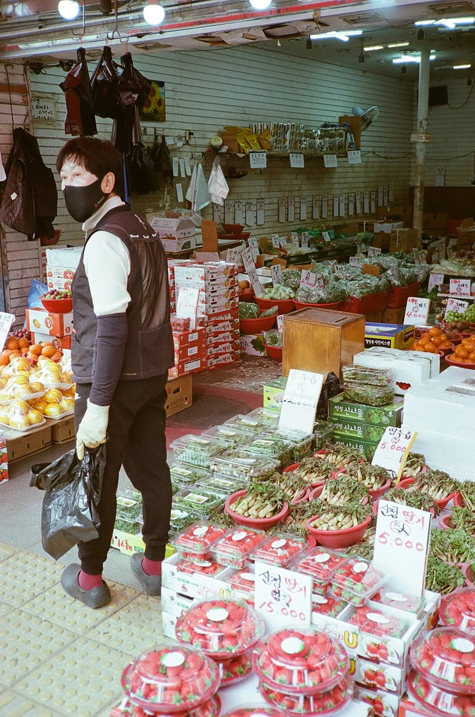 A person wearing a black face mask, white gloves, and black clothing stands in a grocery store near produce and packaged fruits, holding a black plastic bag.