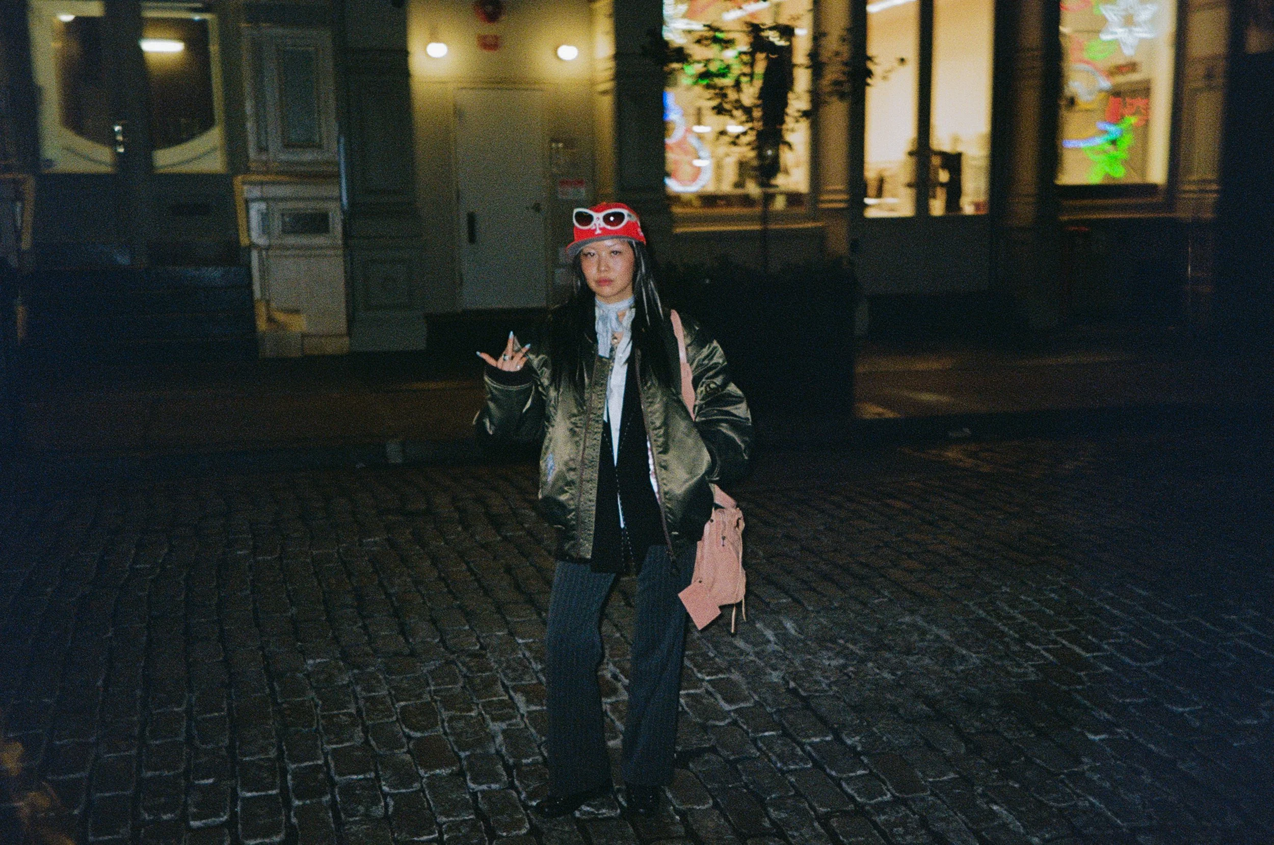 A woman stands on a cobblestone street at night, wearing a brown jacket, pinstripe pants, a red hat with decorative eyes, and holding a handbag, with illuminated storefronts in the background.