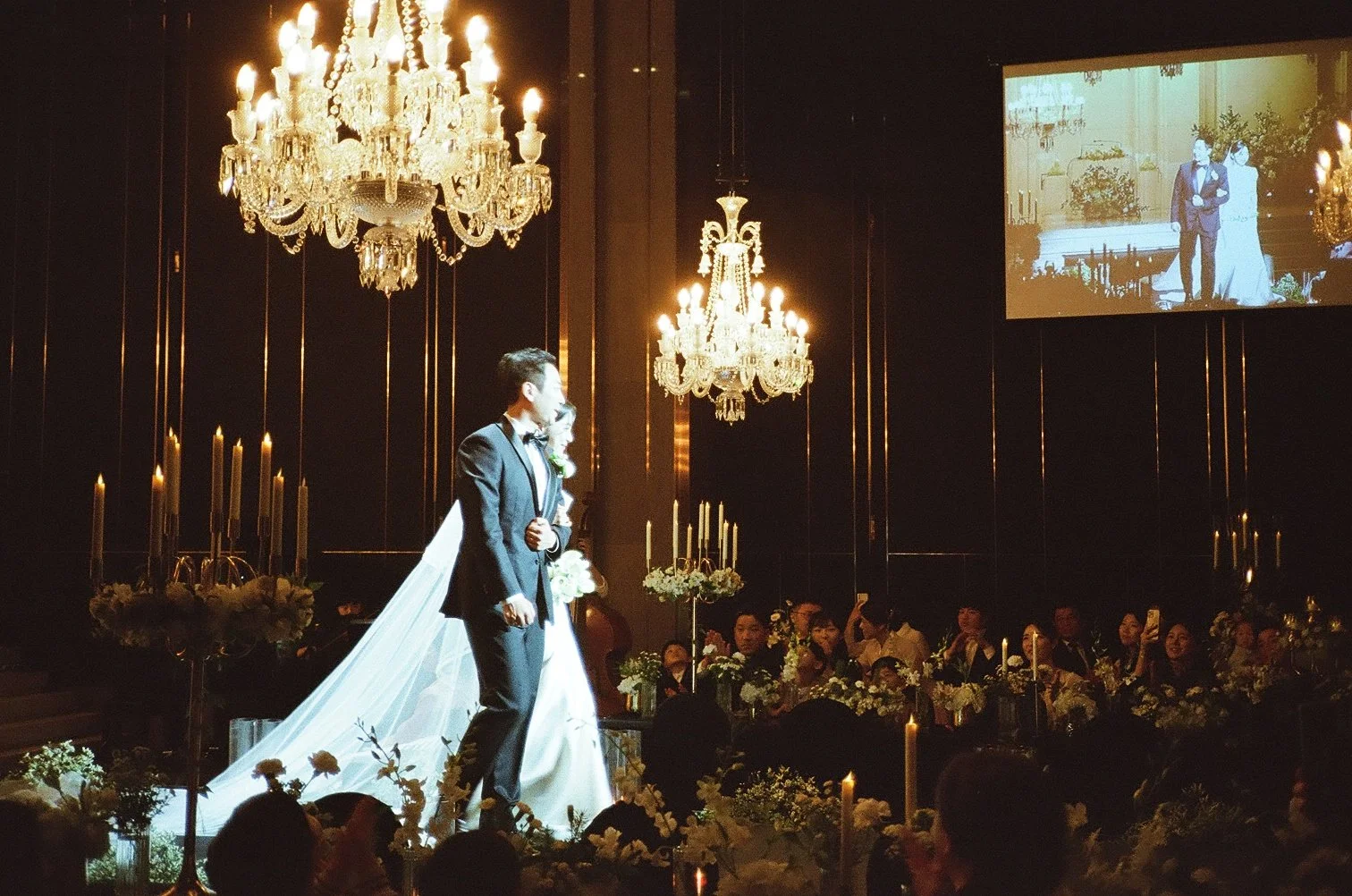 A wedding ceremony with a bride and groom standing under chandeliers and candles, with guests seated at tables and a large screen showing their image.