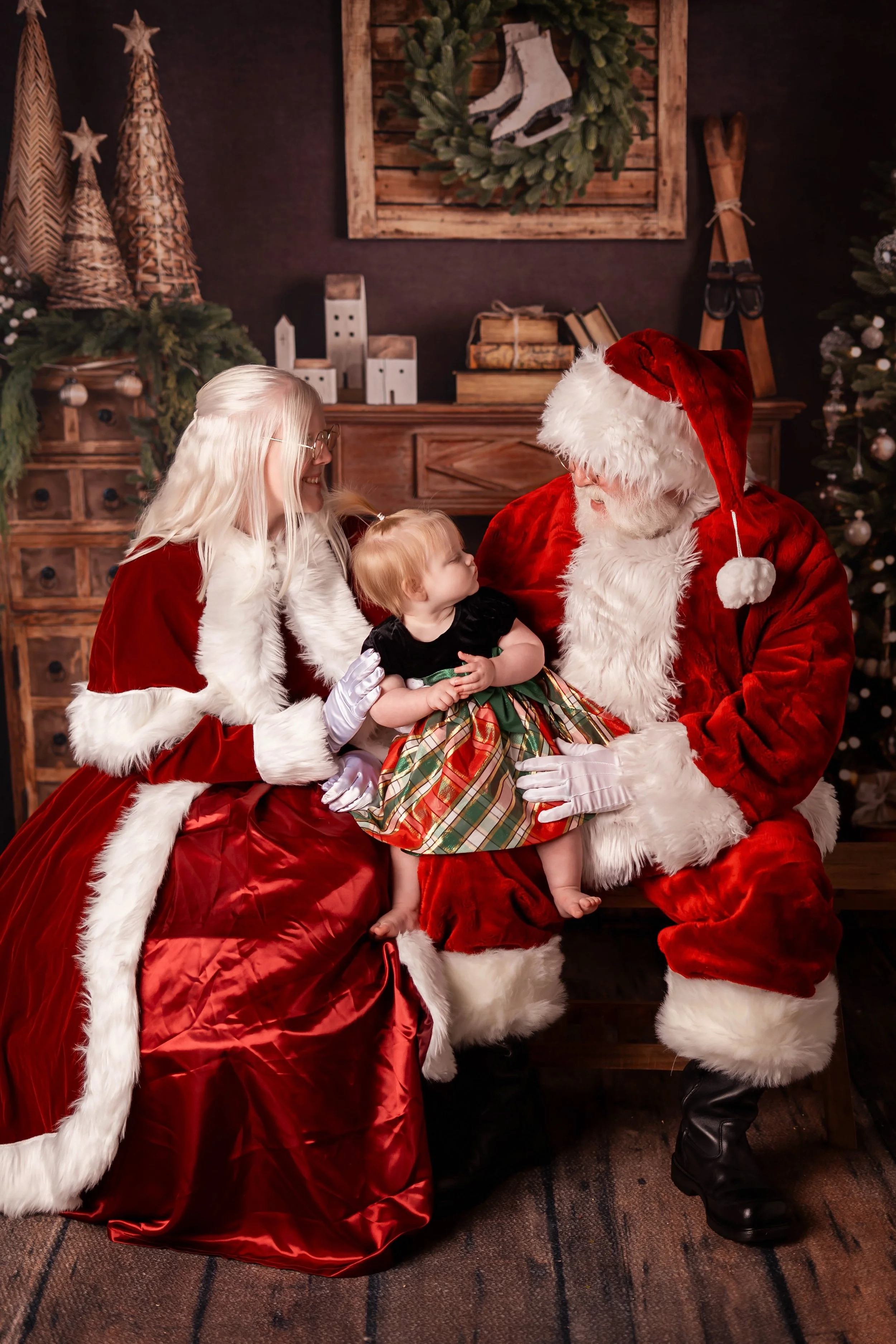 A baby in a Christmas dress sits on Santa Snows lap looking into each others eyes as Mrs. Claus looks at the baby