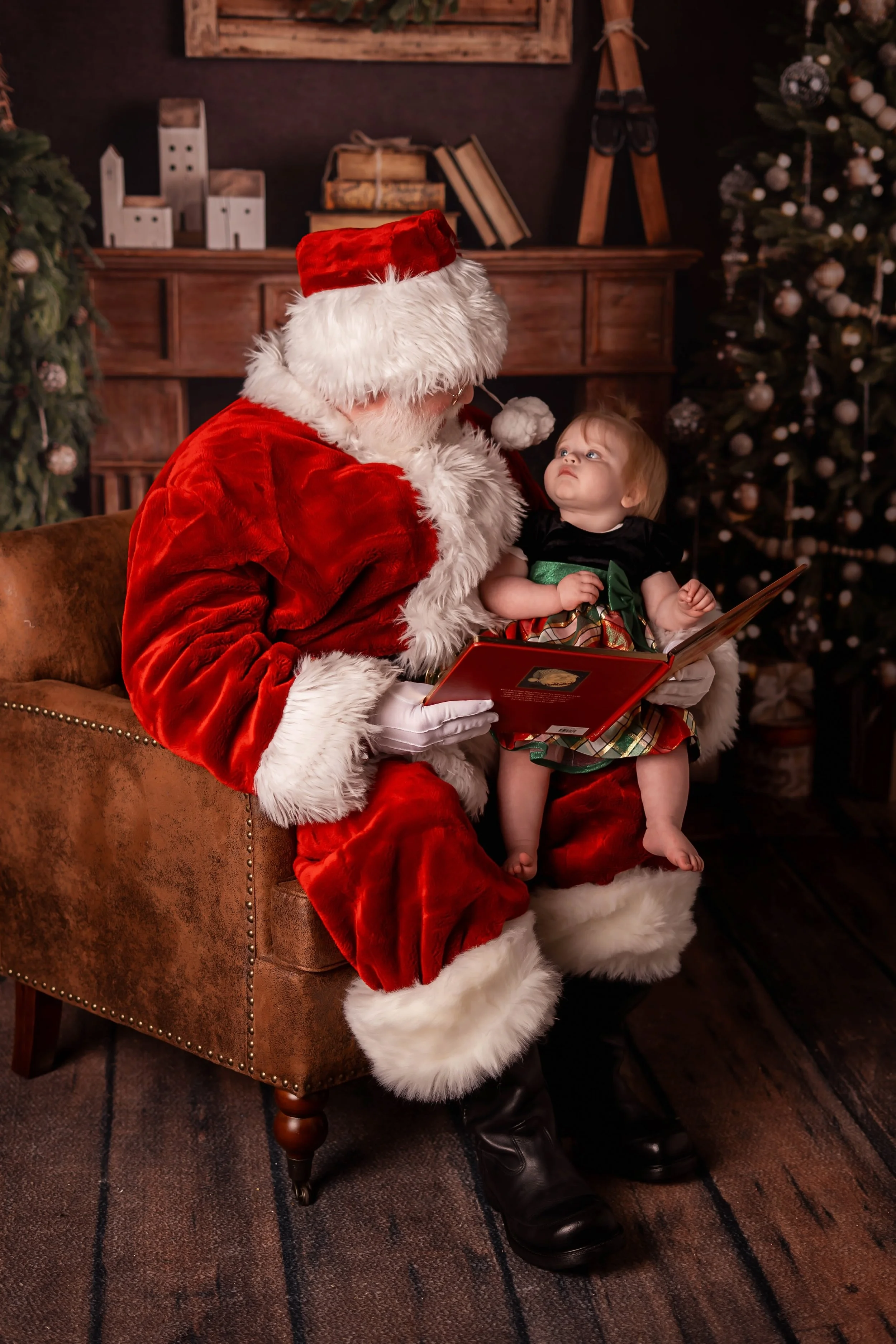 Santa Snow is seated in a chair with a baby on his knee reading The Night Before Christmas as she stairs up at Santa