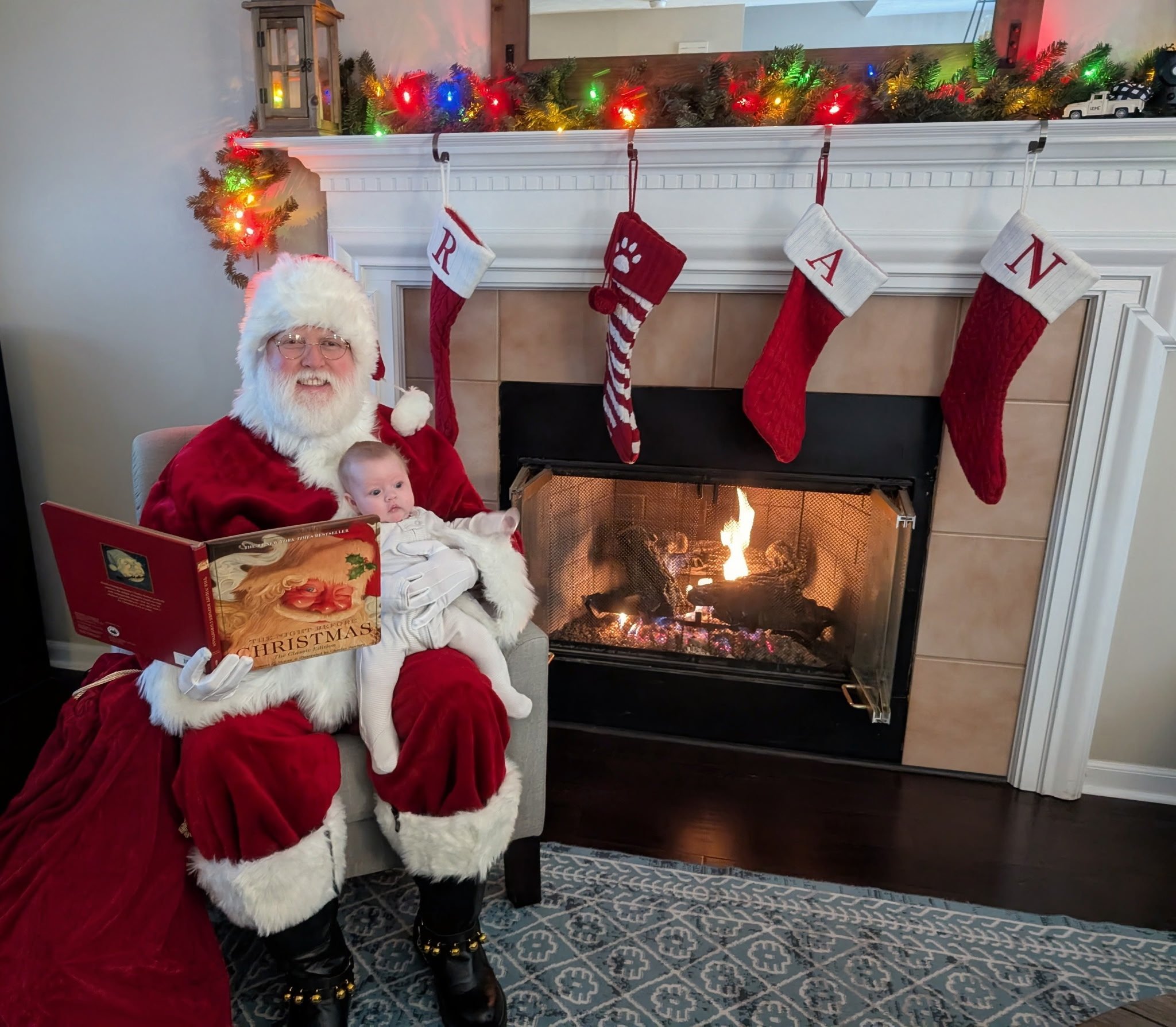 A man dressed as Santa Claus reads a Christmas book to a baby sitting on his lap in front of a lit fireplace decorated with stockings and colorful Christmas lights.