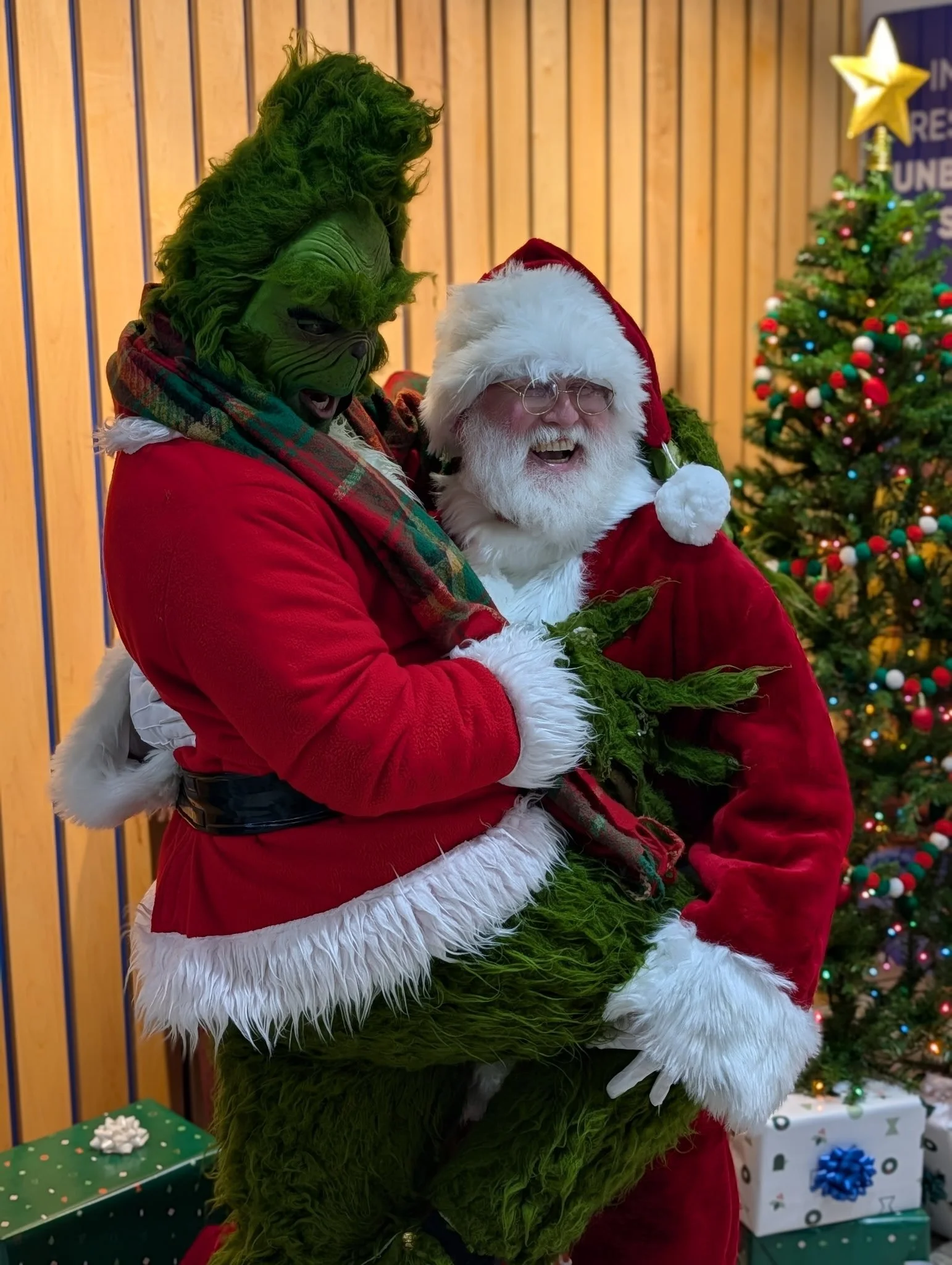Santa Claus laughing while holding a person dressed as the Grinch, who is also laughing, in a festive Christmas setting with decorated Christmas trees and wrapped presents.