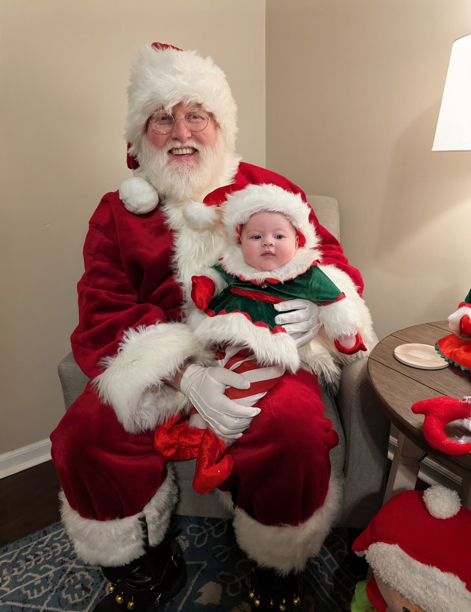 Santa Claus holding a young child dressed in Christmas-themed clothing, sitting in a room decorated for Christmas.