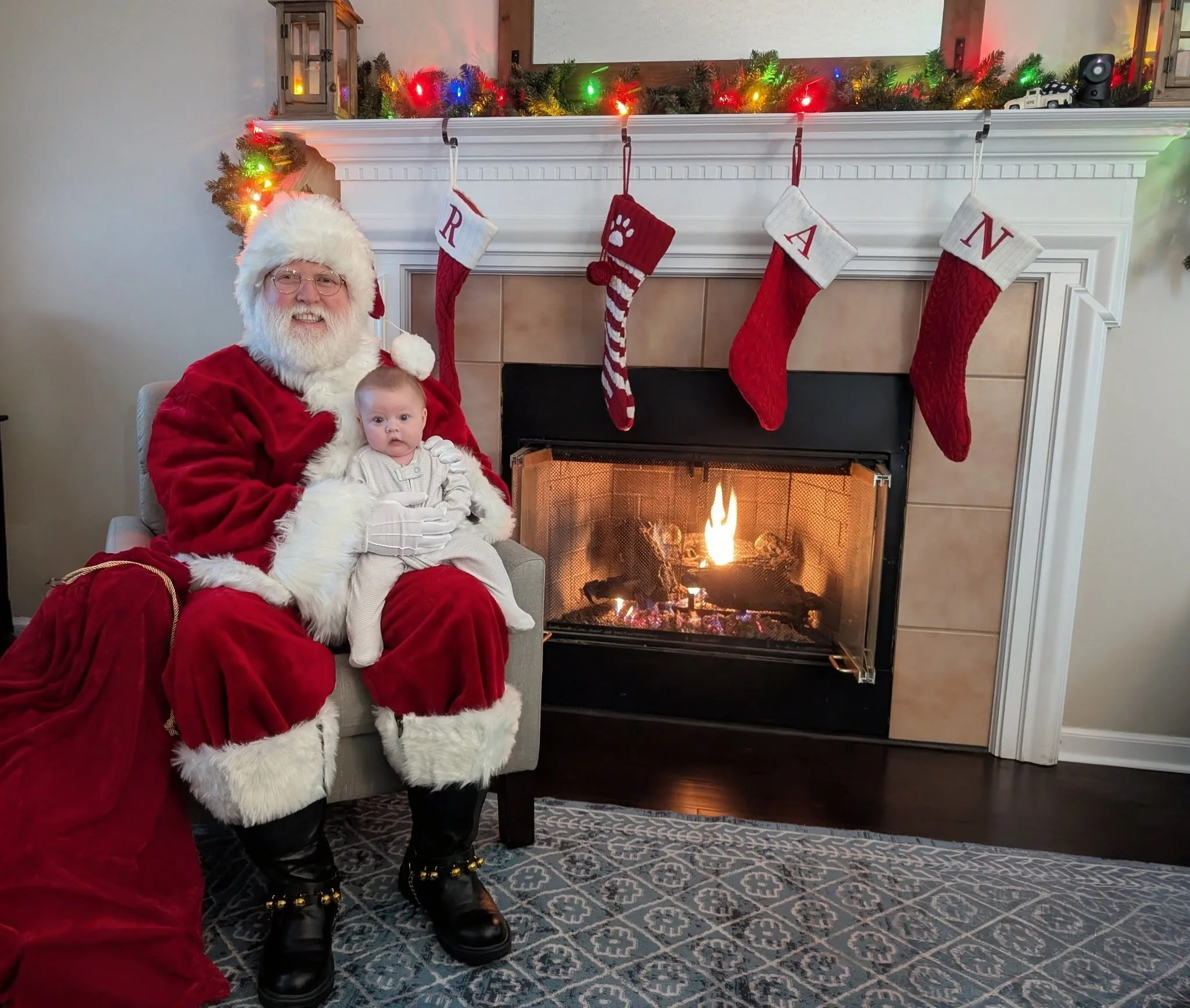 Santa Nick sits by a fire fireplace with a baby on his lap, toy bag on his side.