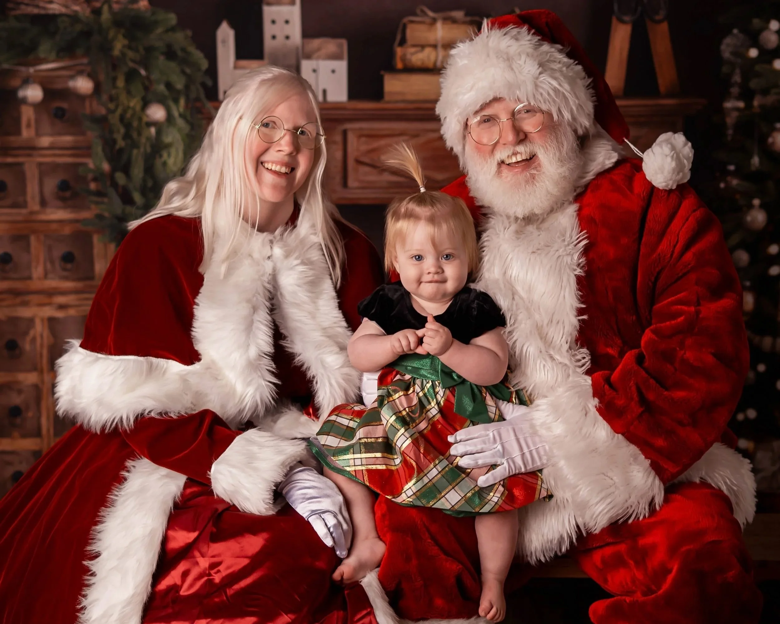 Santa Snow and Mrs. Claus are seated with a baby on their lap smiling at the camera.
