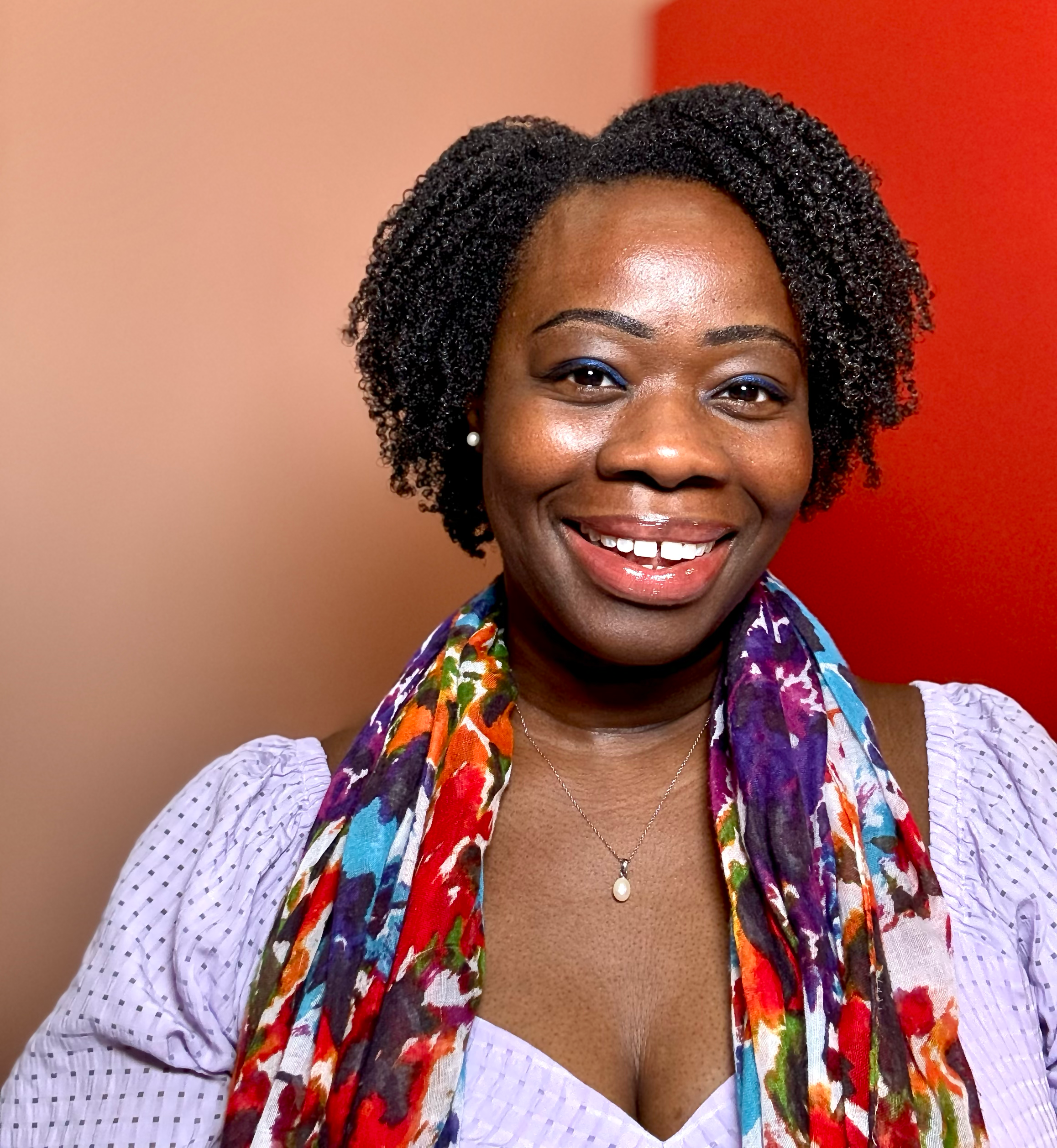 A smiling African American woman with short curly hair, wearing a purple blouse, a floral scarf, and a pearl necklace, standing against a colorful background.