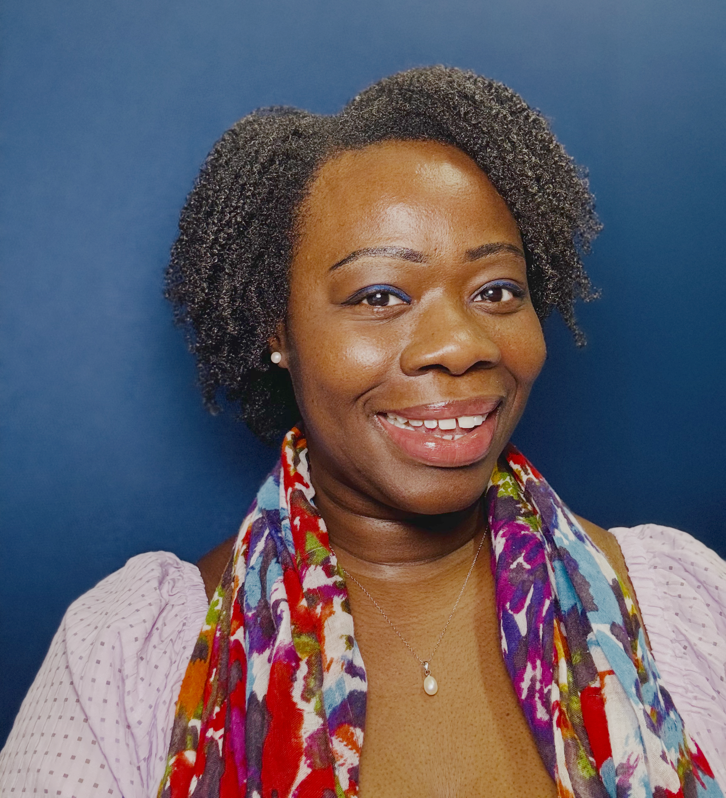 A smiling woman with short, curly hair wearing a colorful scarf, pearl earrings, and a light pink blouse against a blue background.