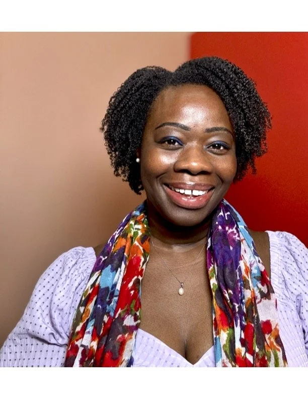 A smiling woman with dark curly hair, wearing a light purple blouse with puffed sleeves and a colorful floral scarf, standing against a neutral background with a red element.