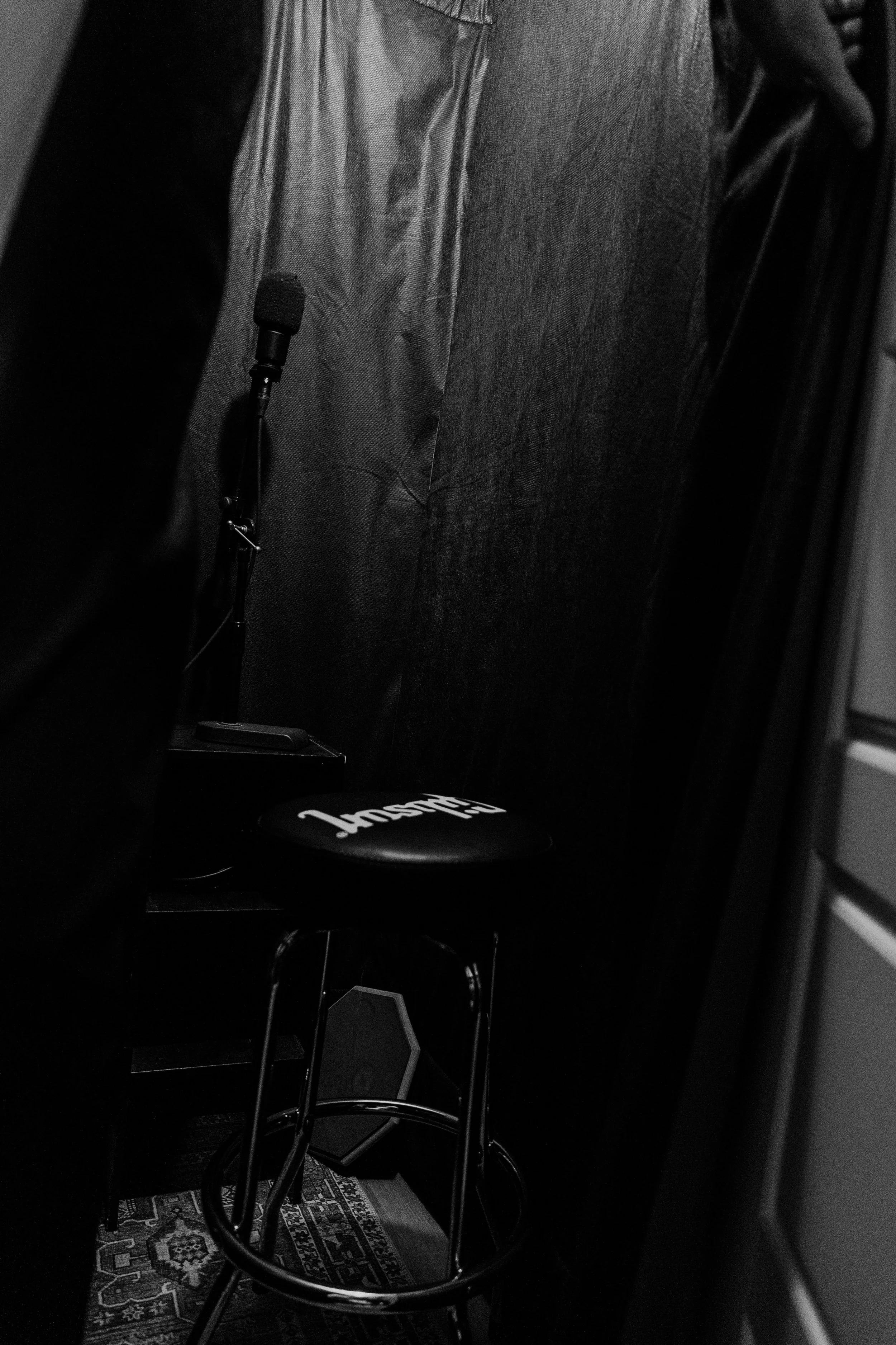 Black stool with the word 'Lush' on the seat, a microphone on a stand, and dark fabric backdrop in a dimly lit room.