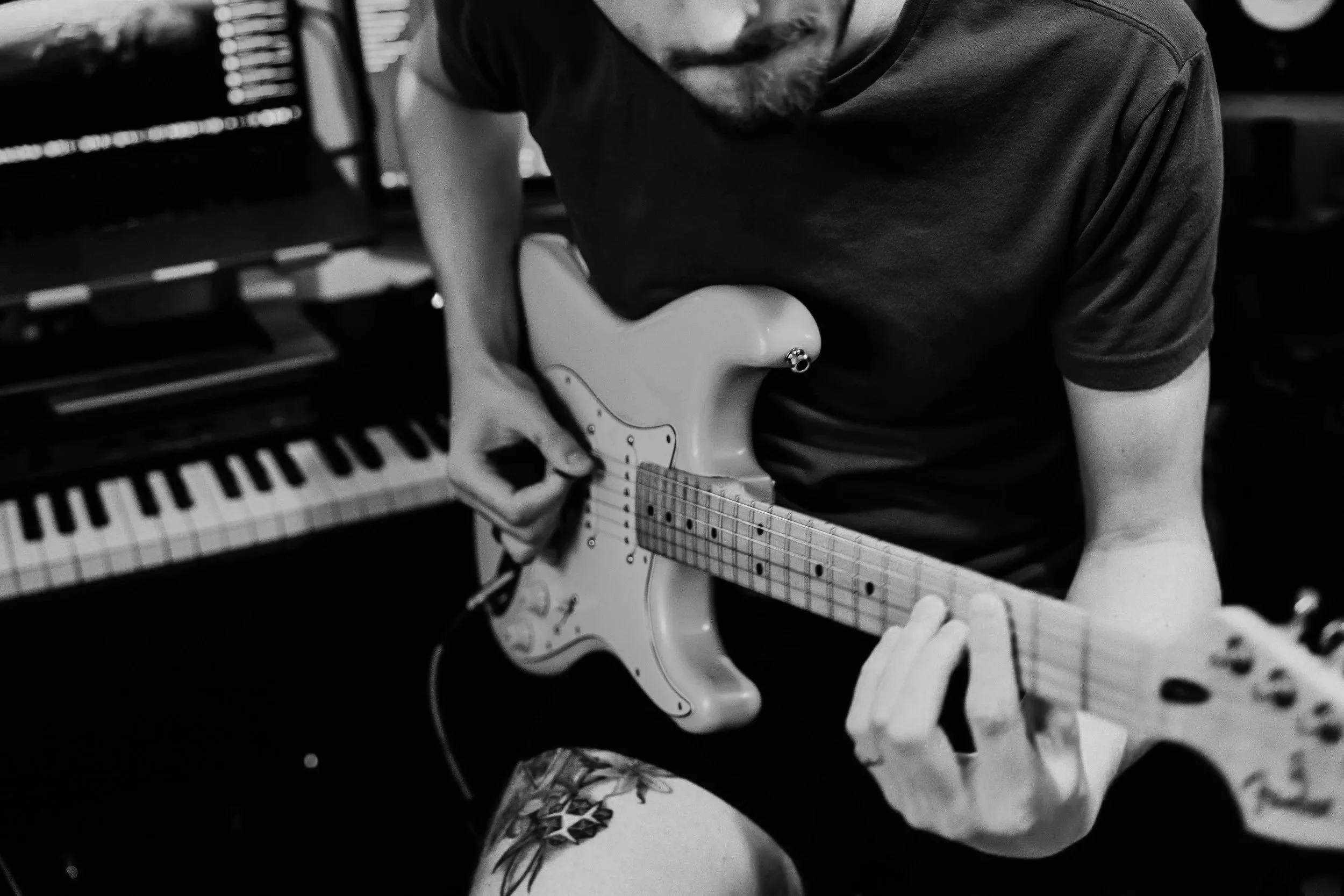 A person playing an electric guitar, sitting in front of a keyboard with a computer monitor in the background. The image is in black and white.