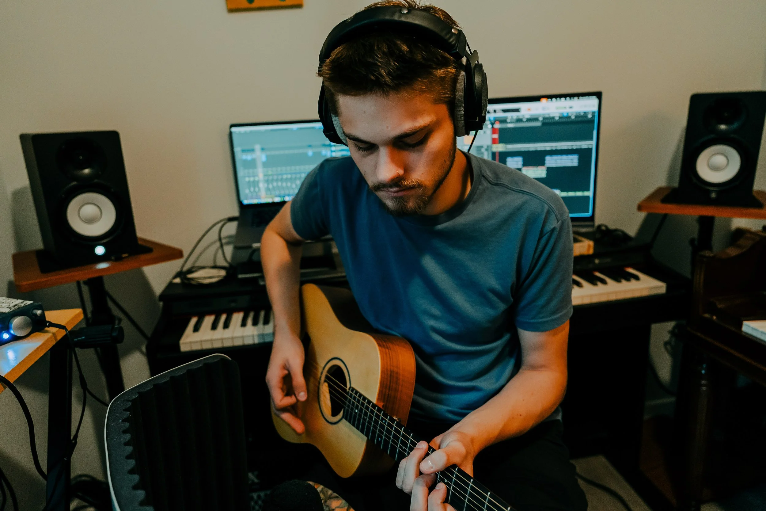 A young man wearing headphones playing an acoustic guitar in a music studio with monitors and electronic equipment.