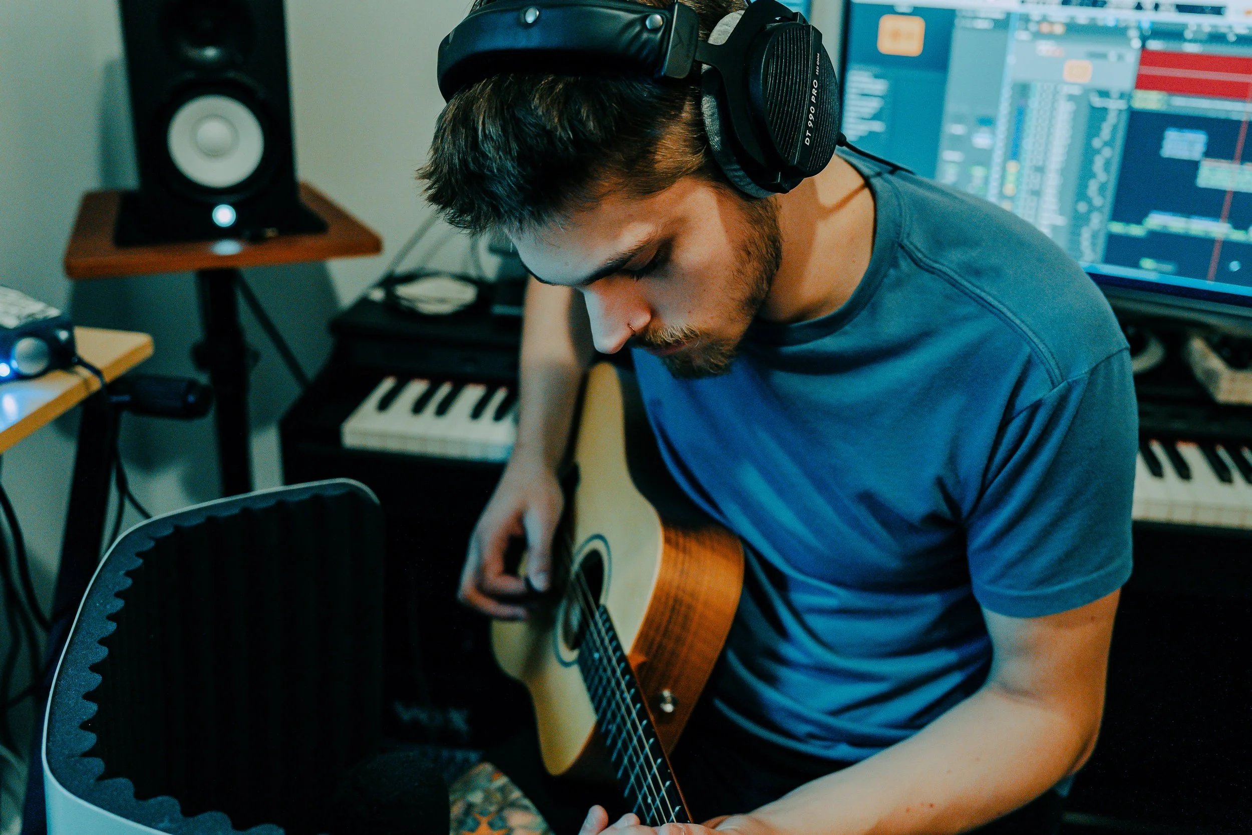 Young man with headphones playing acoustic guitar in a music studio surrounded by audio equipment, keyboard, and computer monitors with music production software.