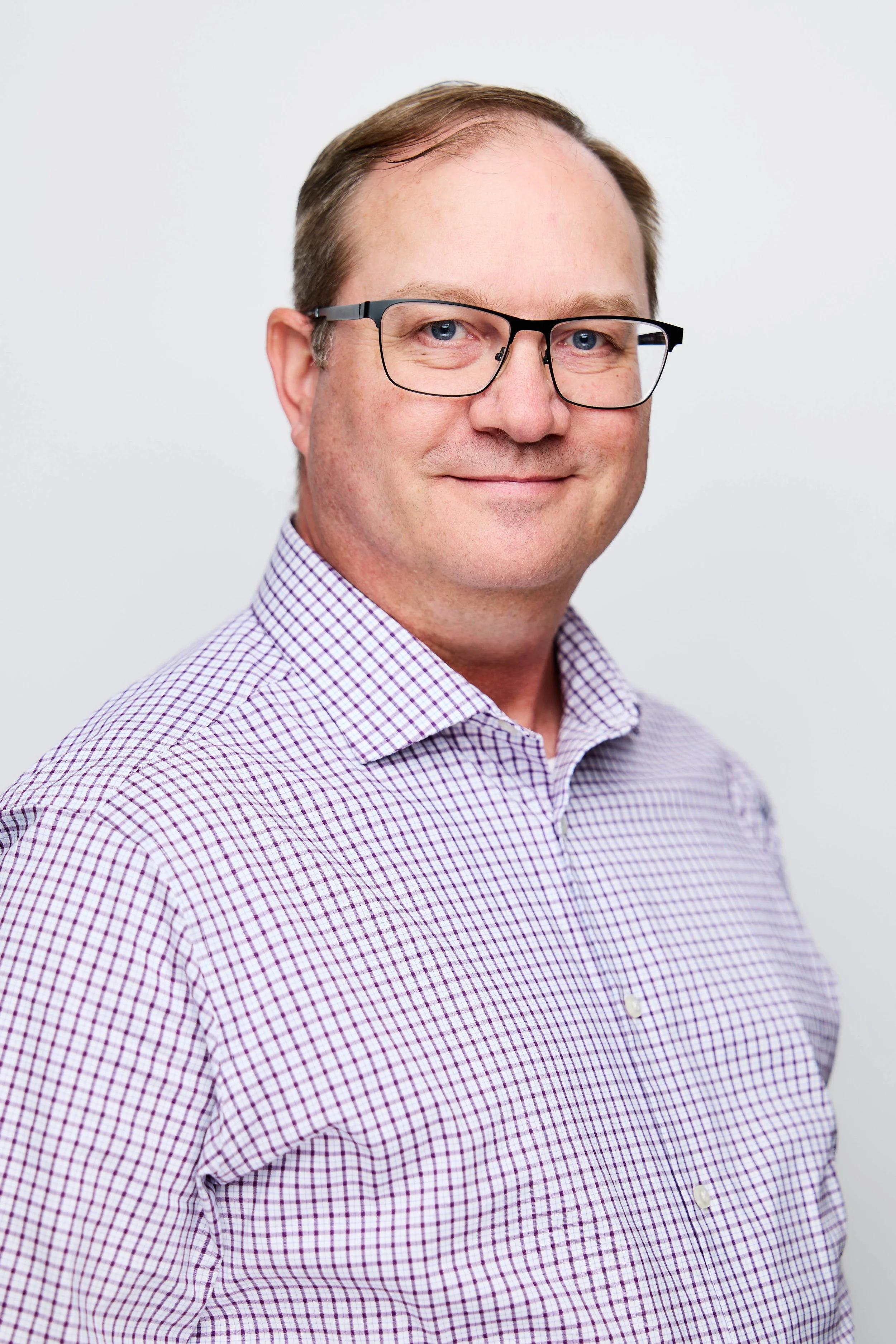 A man wearing glasses and a checkered purple and white shirt, smiling against a plain white background.