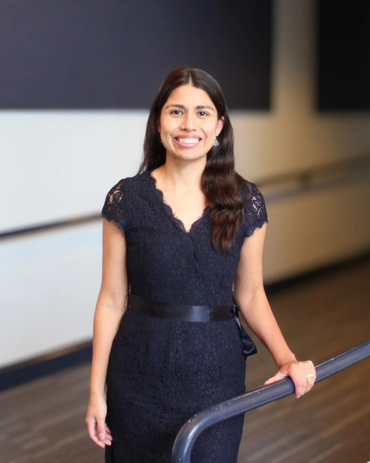 A woman with long dark hair wearing a navy lace dress, smiling, standing by a railing in an indoor setting.