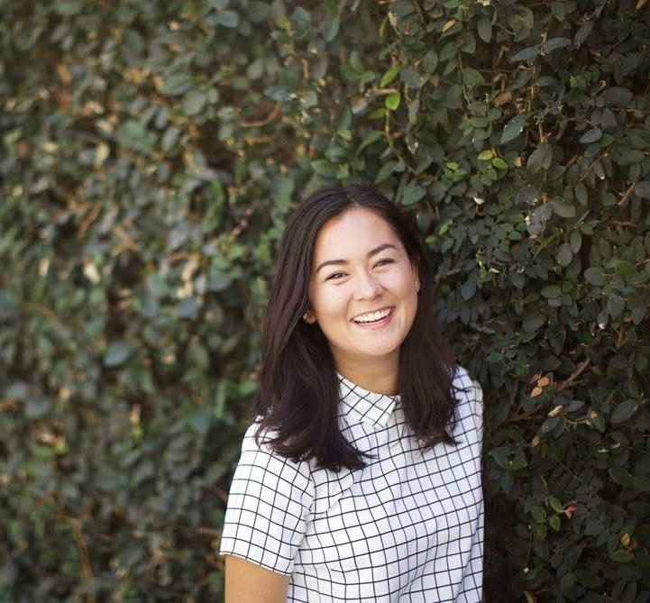 A young woman with long dark hair is smiling and standing outdoors in front of a dense green foliage background, wearing a checked shirt who is a happy client of Making Meaning