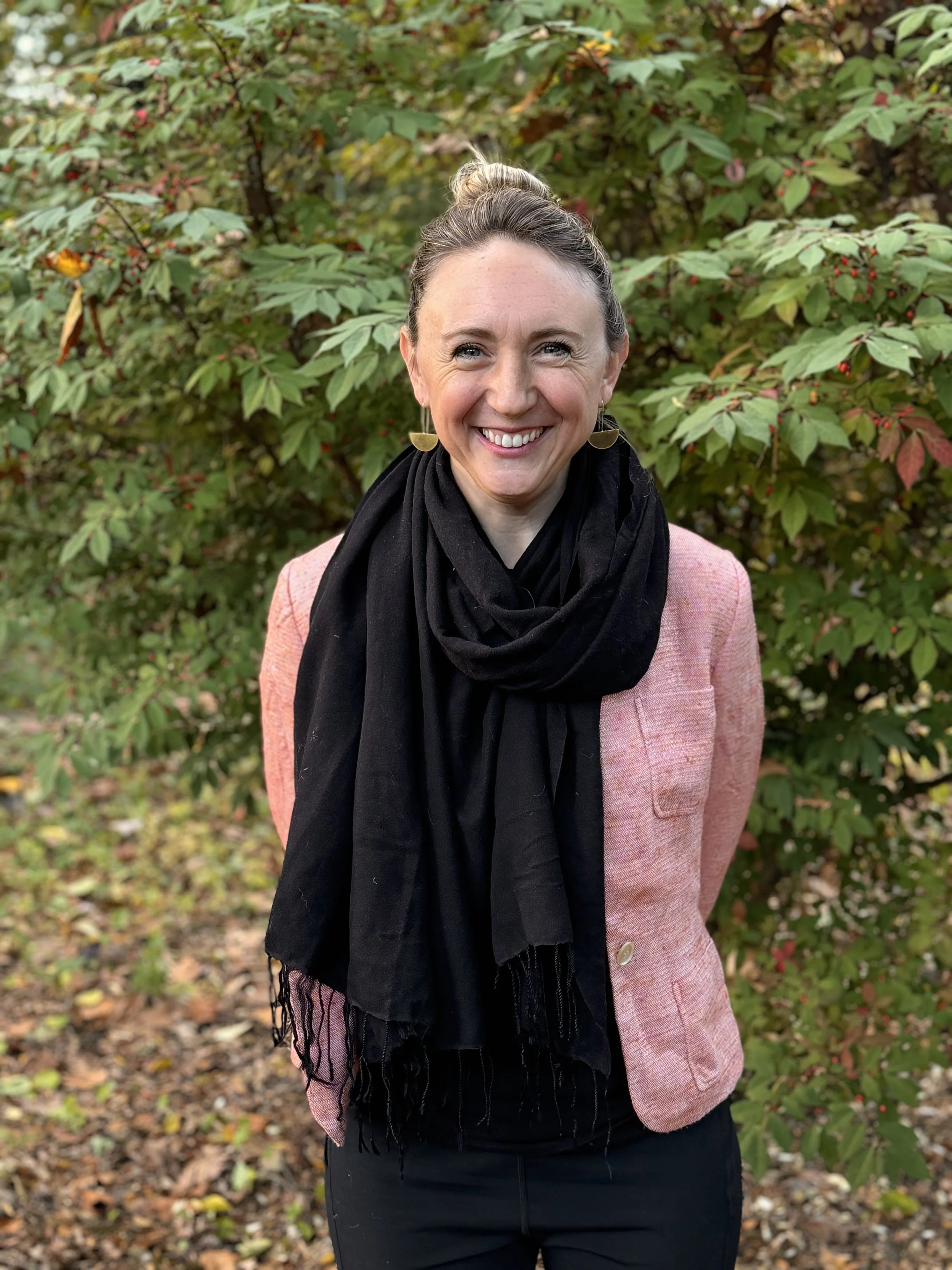 Katy Mixter, Leadership Coach, is smiling outdoors in front of green and red foliage, wearing a pink blazer, black scarf, and black pants.