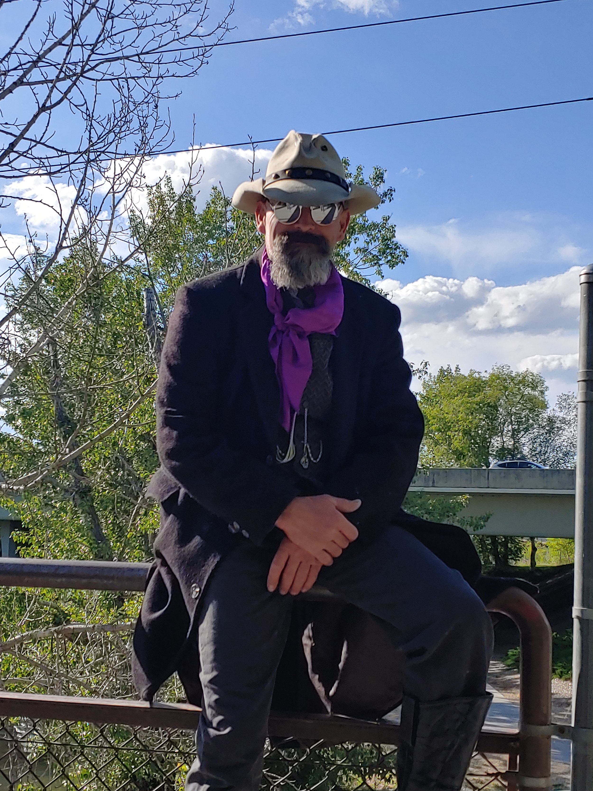 A man with a beard wearing sunglasses, a wide-brimmed hat, a purple bandana, a black blazer, and dark pants sitting on a railing outdoors with trees and a blue sky in the background.