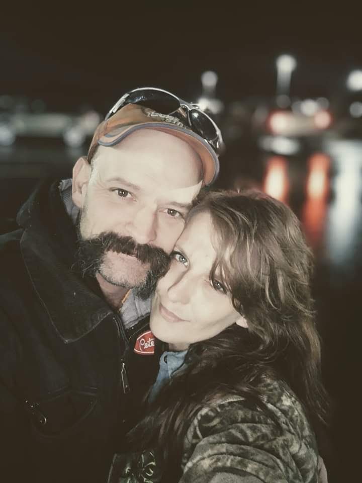 A man with a beard and mustache, wearing a cap and sunglasses, and a woman with wavy hair, are close together outdoors at night, with blurred lights and water reflections in the background.
