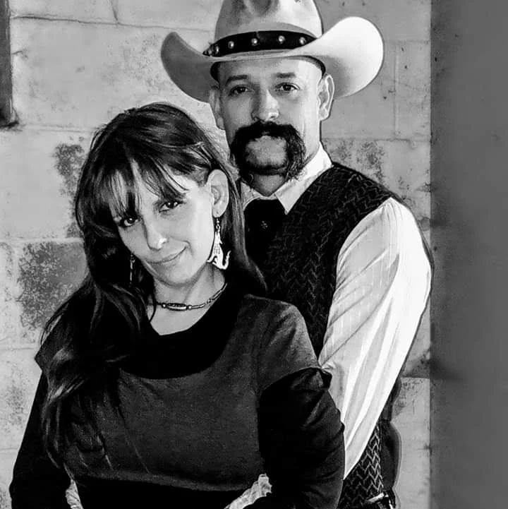 A black and white photo of a man in a cowboy hat and a woman with long dark hair, smiling at the camera, standing close together against a brick wall.