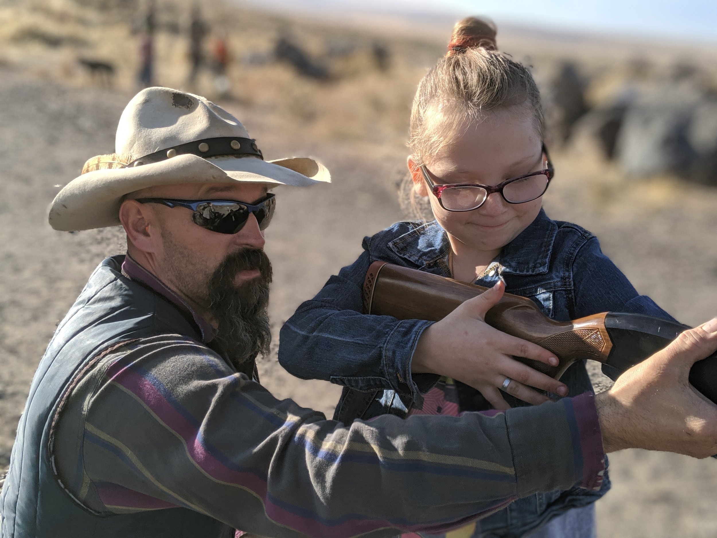 A man with a beard, wearing a cowboy hat, sunglasses, and a plaid shirt, assisting a young girl with glasses and a denim jacket in aiming a rifle outdoors on a sunny day.