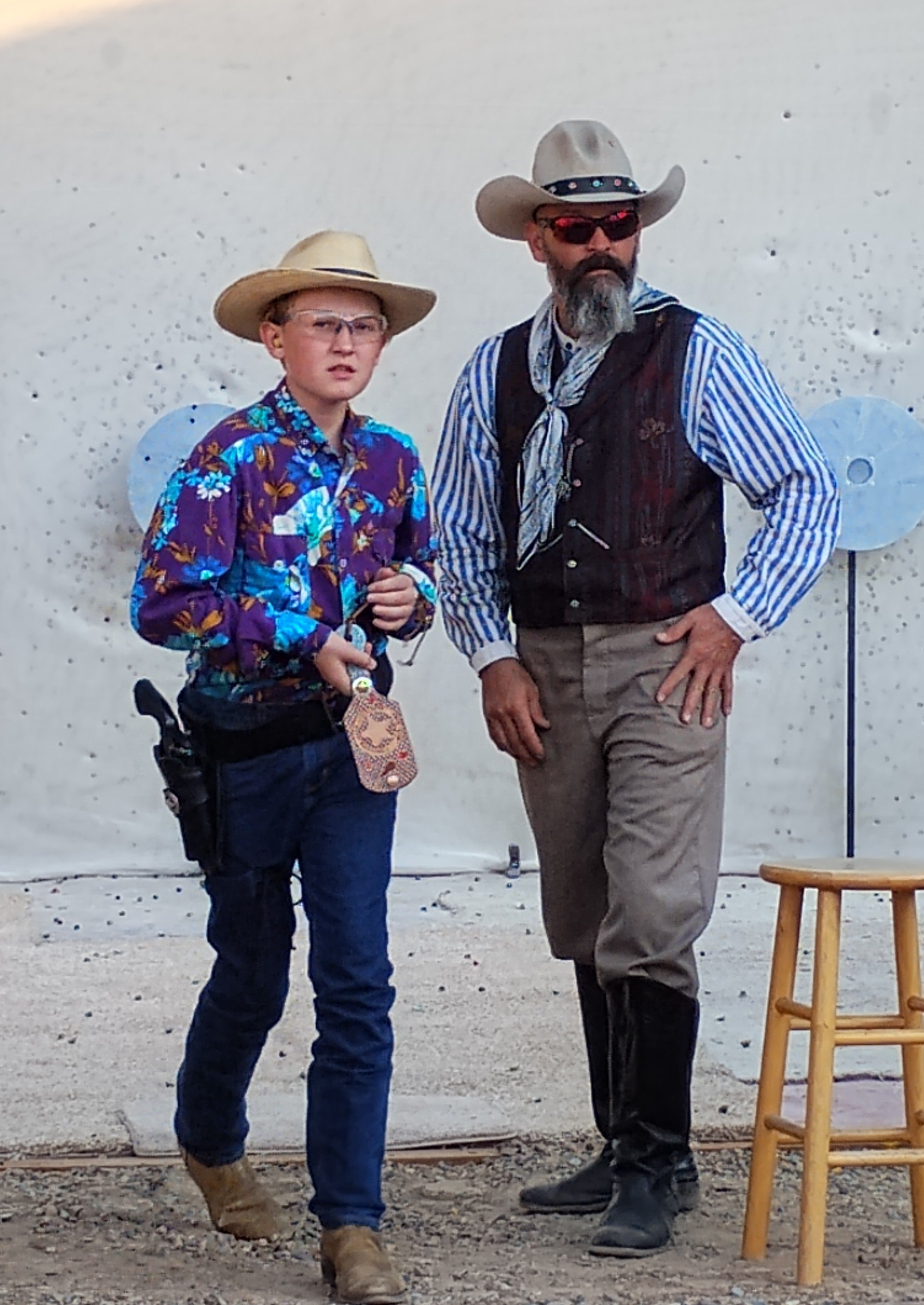 Two men dressed as cowboys standing in front of a white wall with target shapes. One younger man is wearing a wide-brimmed hat, glasses, a colorful Hawaiian shirt, jeans, and boots, holding a lanyard and a small pouch. The older man is wearing a cowboy hat, sunglasses, a striped shirt, a vest, khaki pants, and black boots, with one hand on his hip.