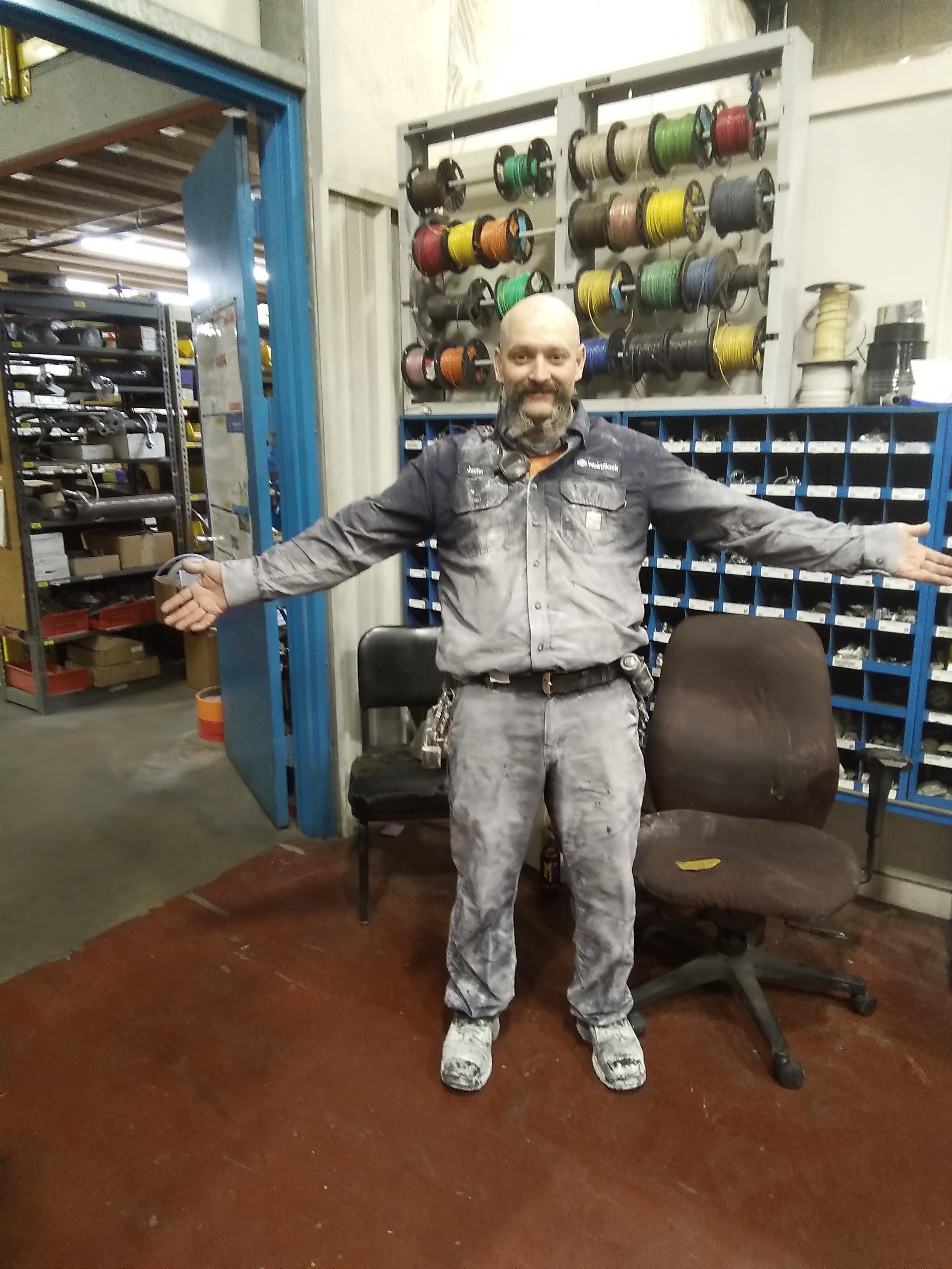 A man with a beard standing in an industrial workshop, wearing work clothes covered in dust, with arms outstretched, surrounded by shelves with wires and tools.