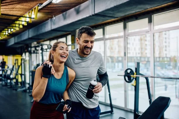 A man and woman smiling and laughing in a gym. The woman wears a blue tank top and gloves, holding a water bottle, while the man wears a gray shirt and holds a phone and water bottle. Large windows and gym equipment are in the background.