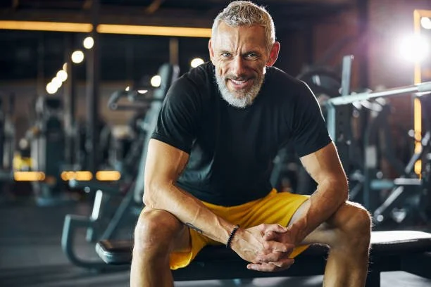 An elderly man with gray hair and beard smiling while sitting on a bench in a gym, surrounded by exercise equipment.