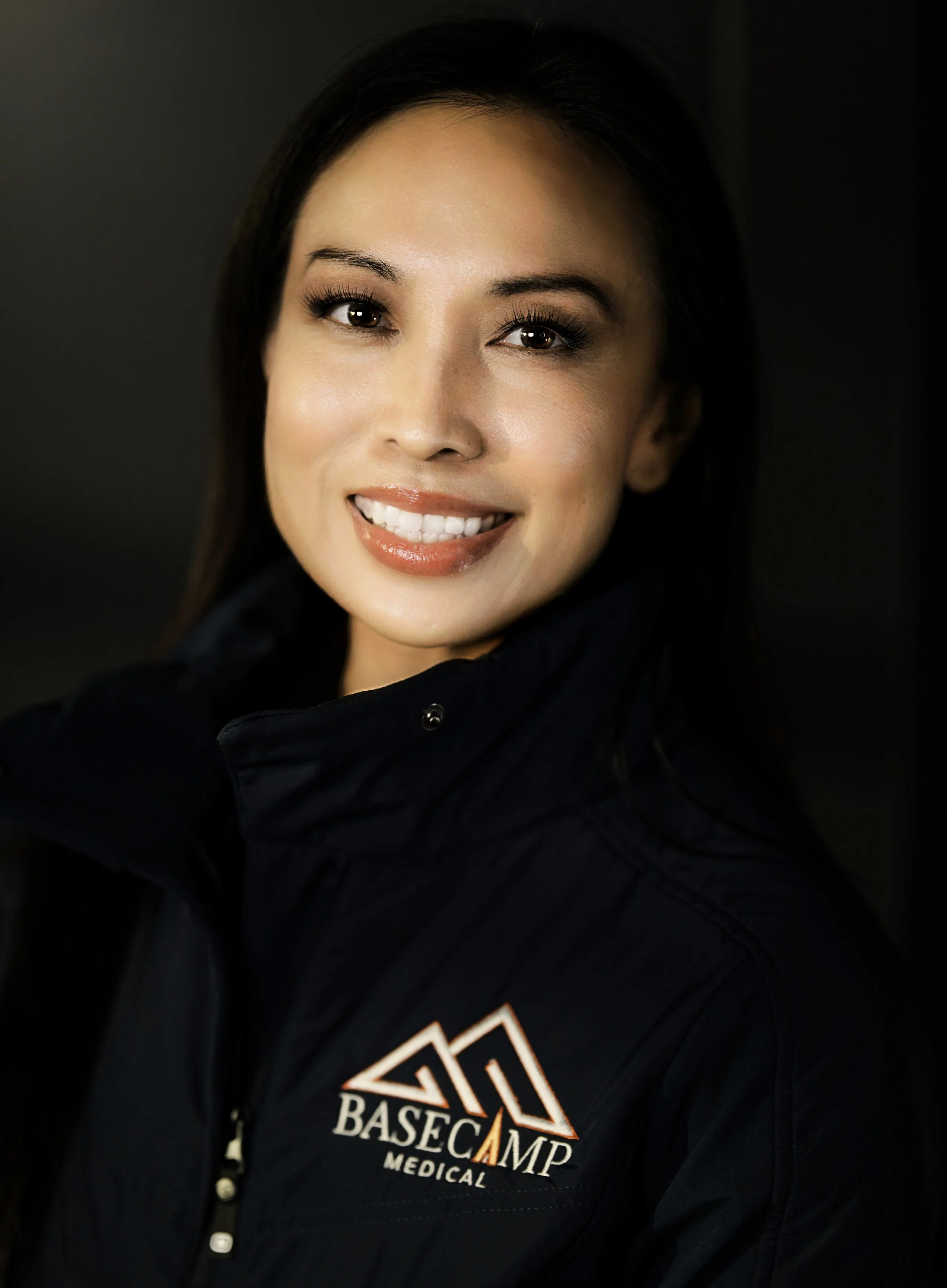 A smiling woman with dark brown hair wearing a black jacket with a logo that reads 'BASE CAMP MEDICAL' on her left side.
