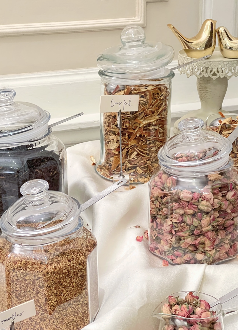 Glass jars filled with dried flowers and herbs on a table, labeled with handwritten tags, part of a decorative display.