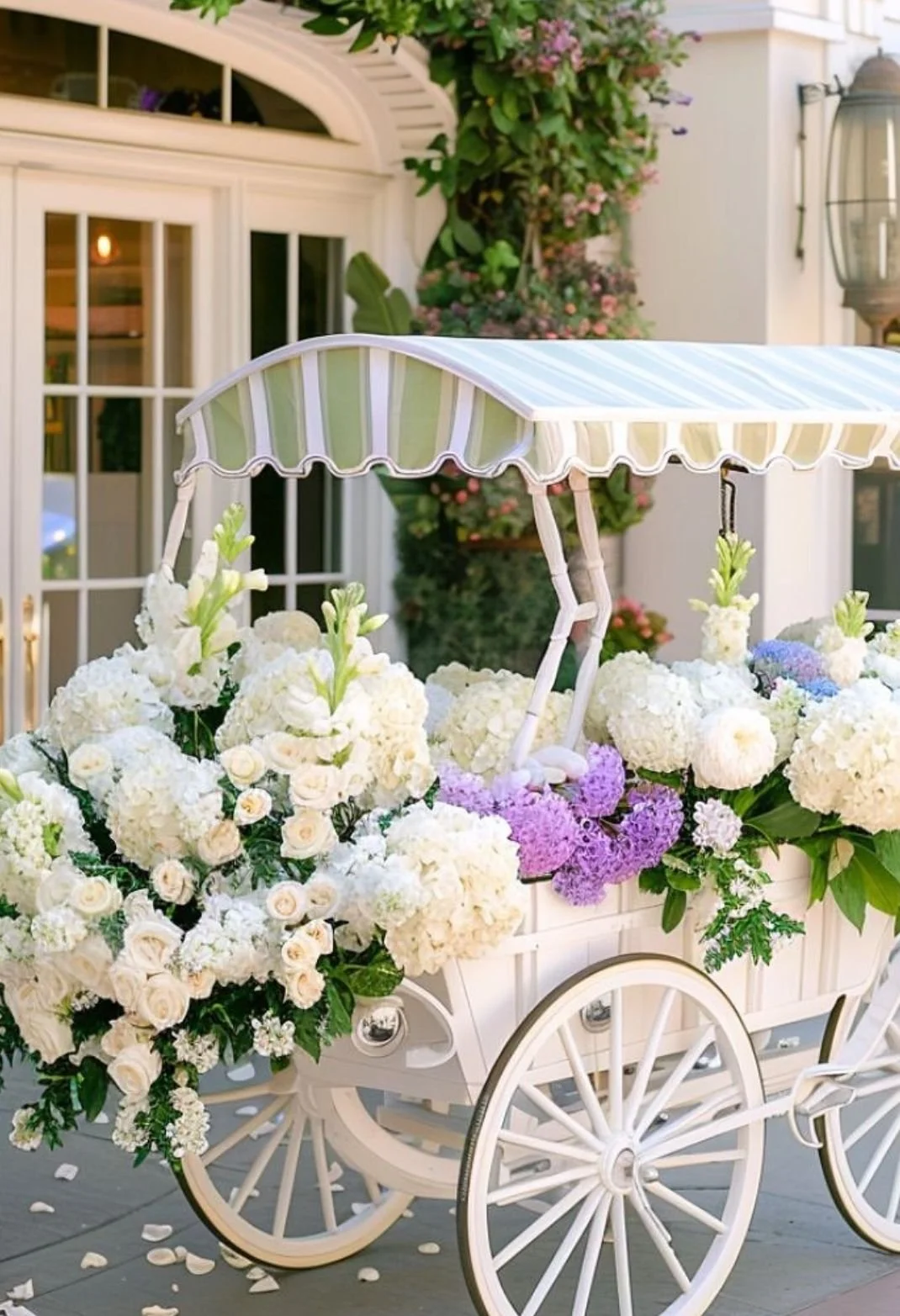 White flower cart with white and purple flowers and a striped green and white canopy, in front of a house with large windows and a lantern