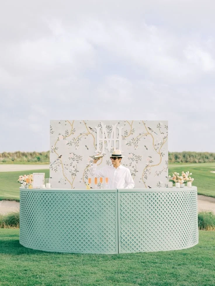 A person wearing a white shirt, sunglasses, and a straw hat standing behind a teal bar outdoors, pouring drinks with a decorative backdrop featuring a tree branch pattern, set on grass with a golf course and cloudy sky in the background.